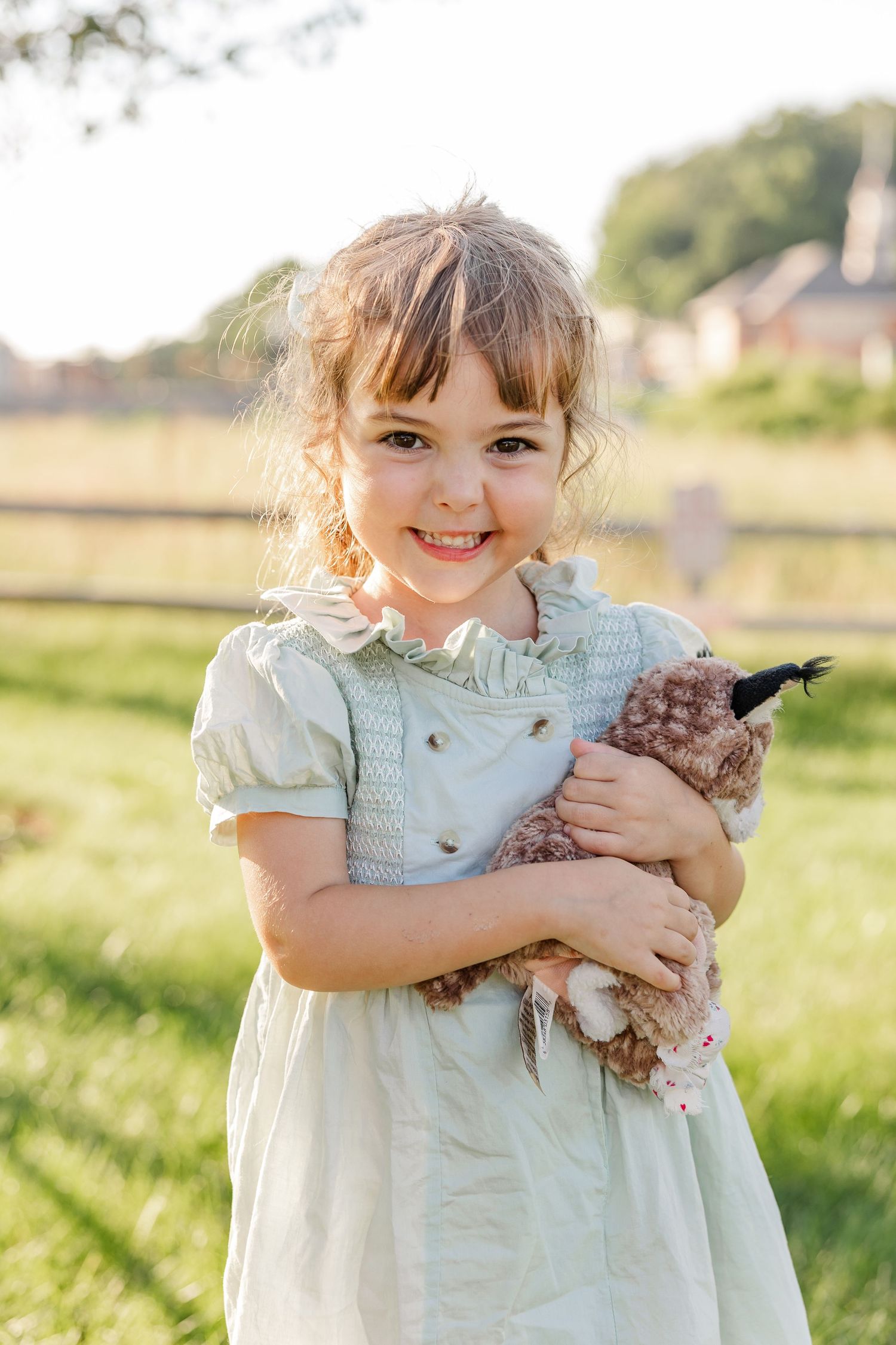 Young girl in mint dress smiles while hugging a stuffed animal toy outdoors in sunny green field.