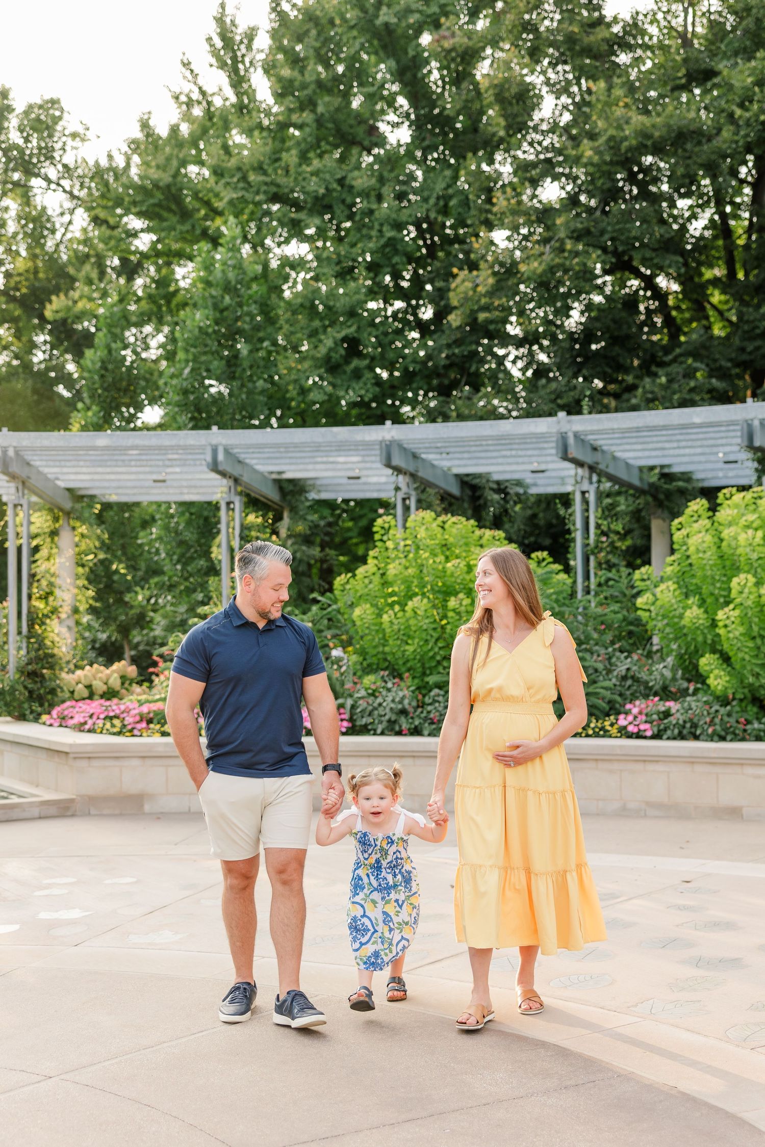 Pregnant woman in yellow dress and husband walk hand in hand with their toddler daughter in a sunny garden.