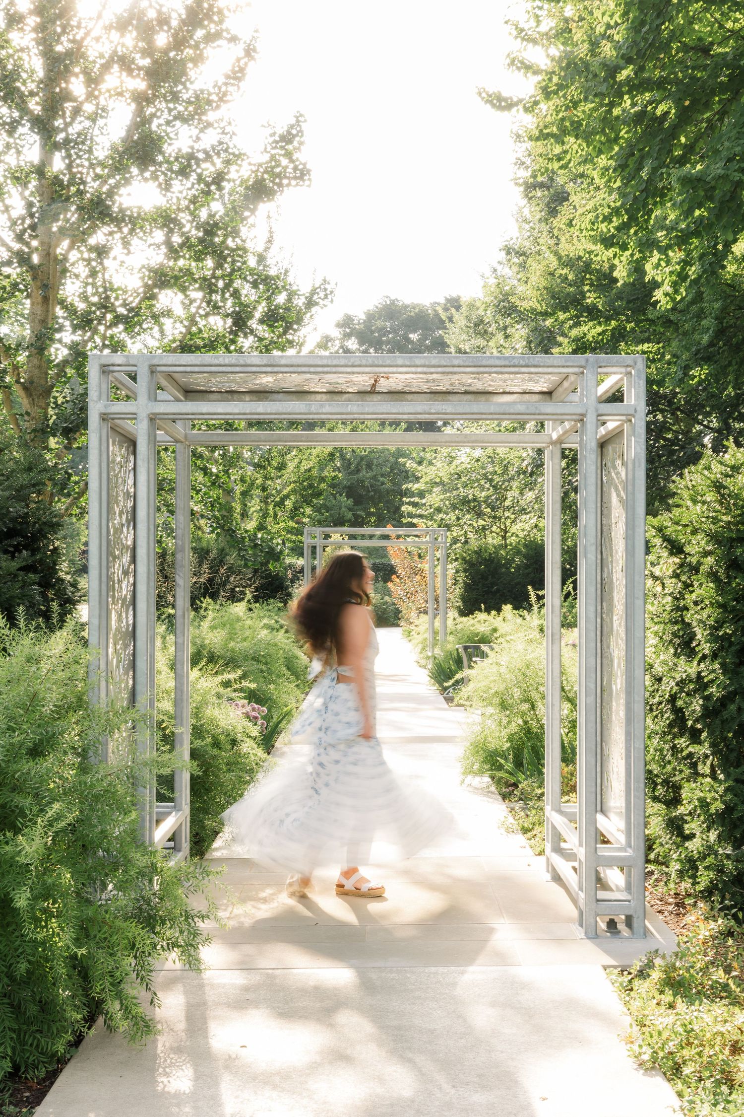 Woman in white dress twirling under garden pergola archway surrounded by lush green trees and plants.