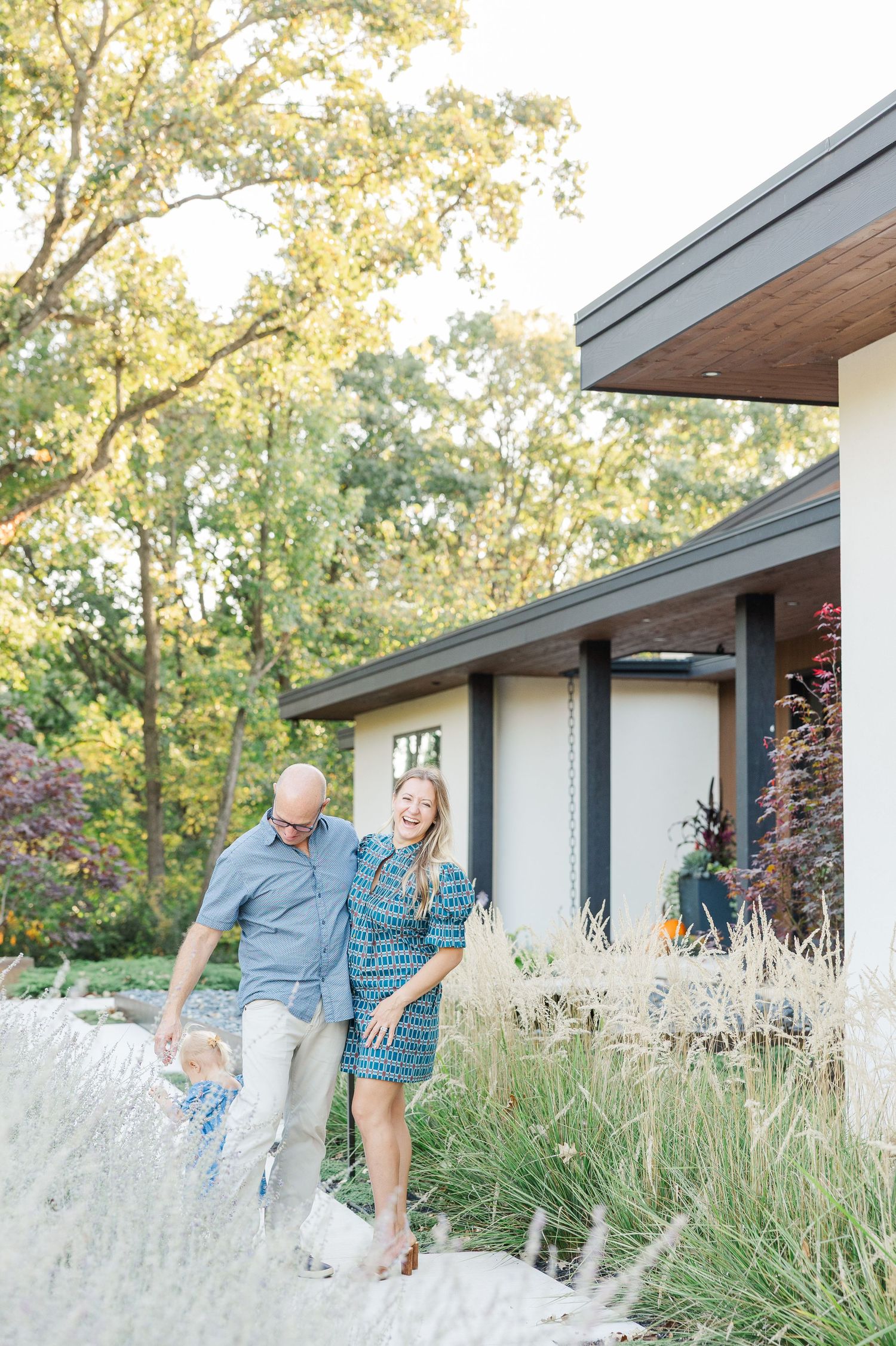 A couple shares playful moments in front of a modern house with ornamental grasses in the foreground.