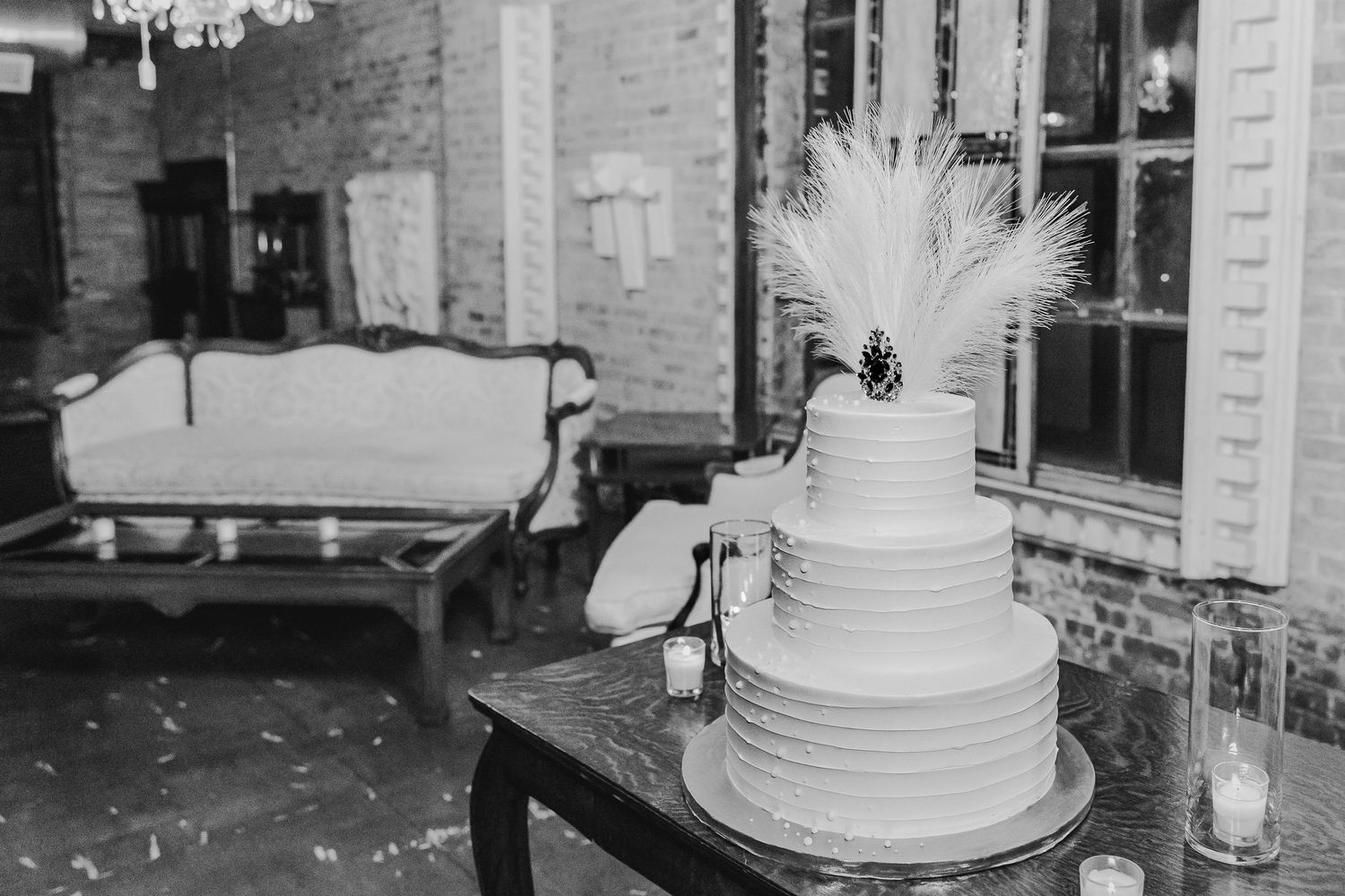 Black and white photo of tiered white wedding cake with feather accent in rustic venue.