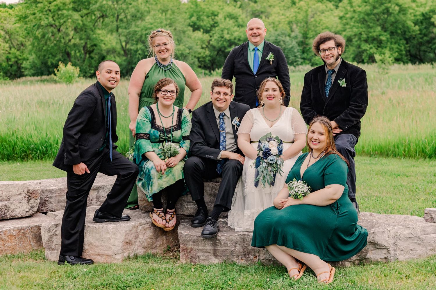 Wedding party in formal attire poses together on stone steps in a green field setting.