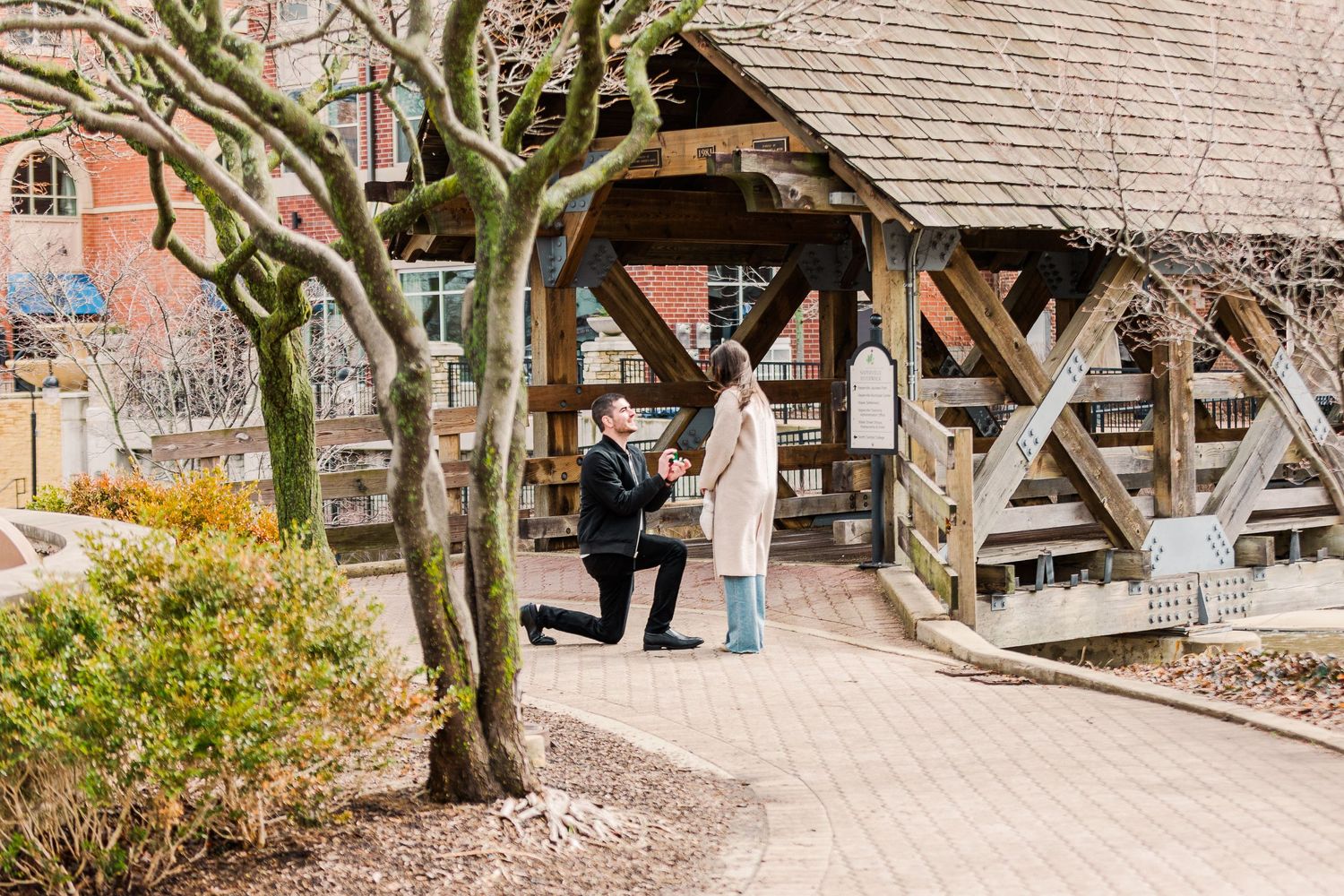 Two people chat under a wooden covered bridge structure surrounded by bare trees.