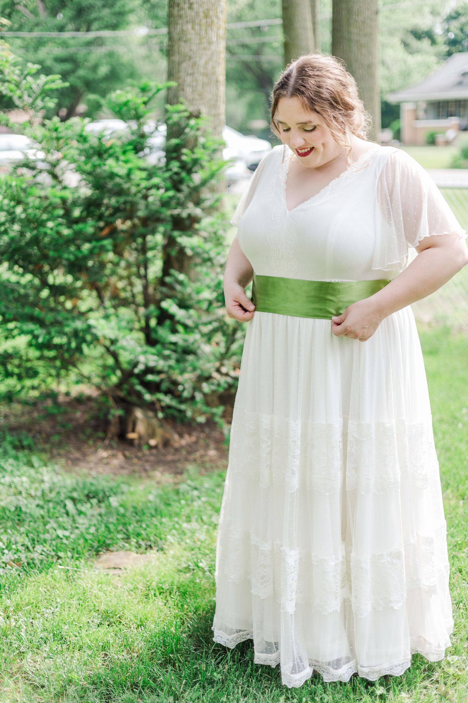 Person in white wedding dress with green sash looks down while adjusting outfit outdoors.