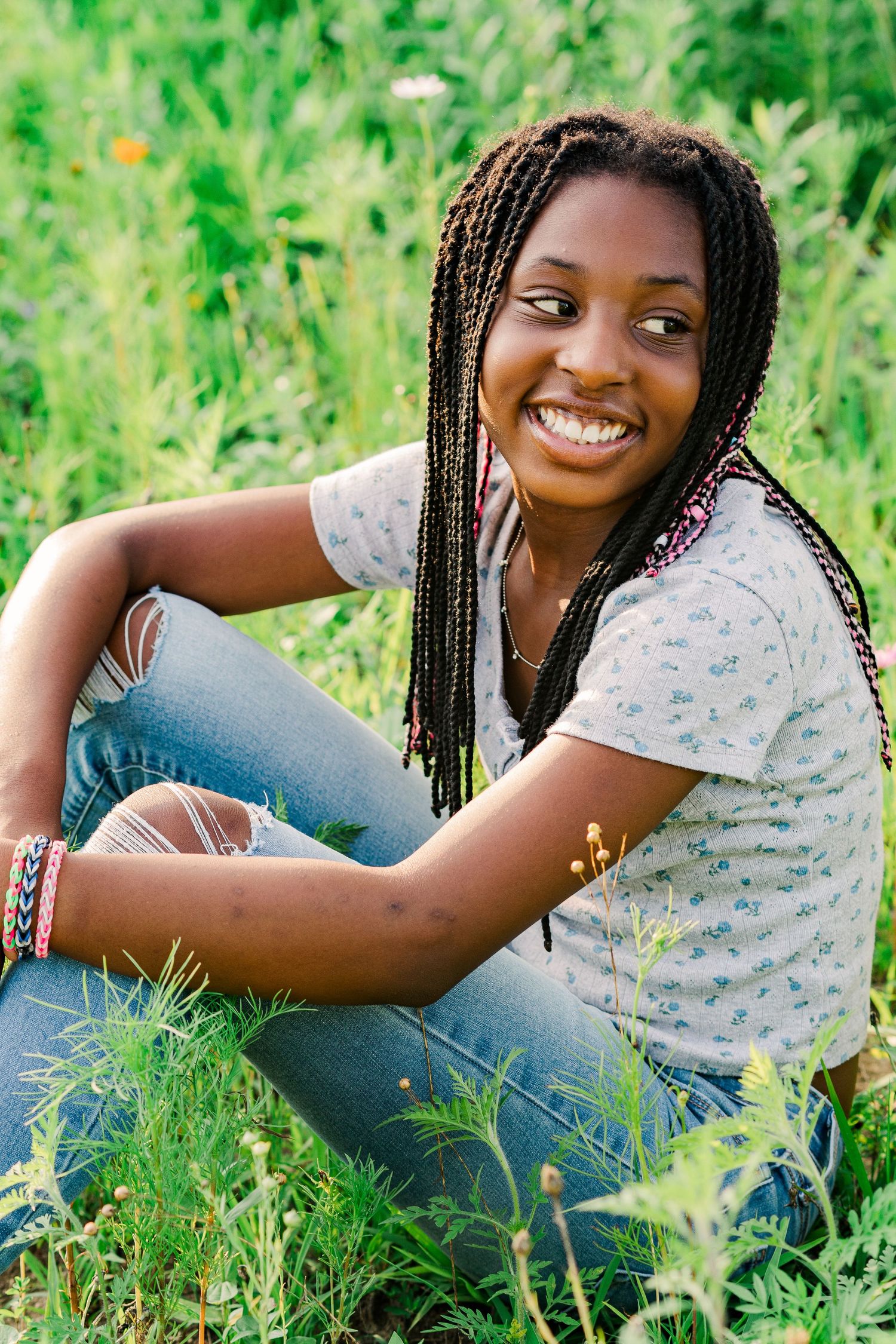 Young woman with long braids smiling while sitting in green grass outdoors.