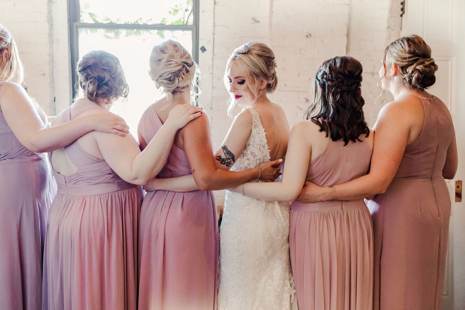 Candid moment of bridal party in dusty rose dresses standing together by sunlit window.