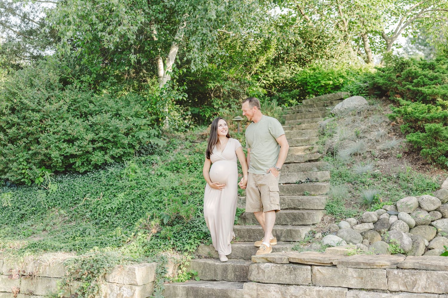 A couple in light casual attire walks up rustic stone steps surrounded by lush green foliage and natural landscaping.