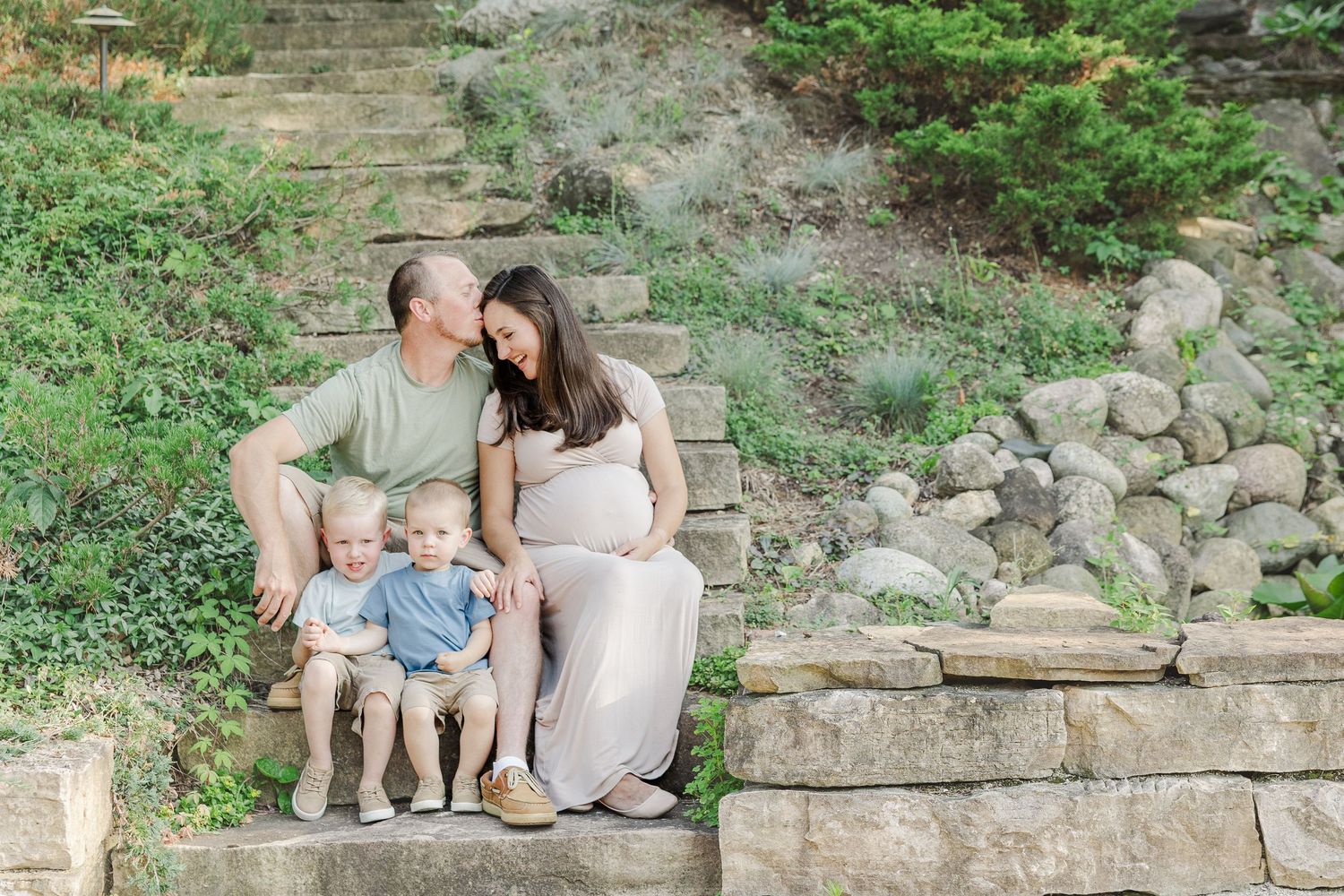 A family sits together on stone steps outdoors in a serene nature setting during a maternity photo session.