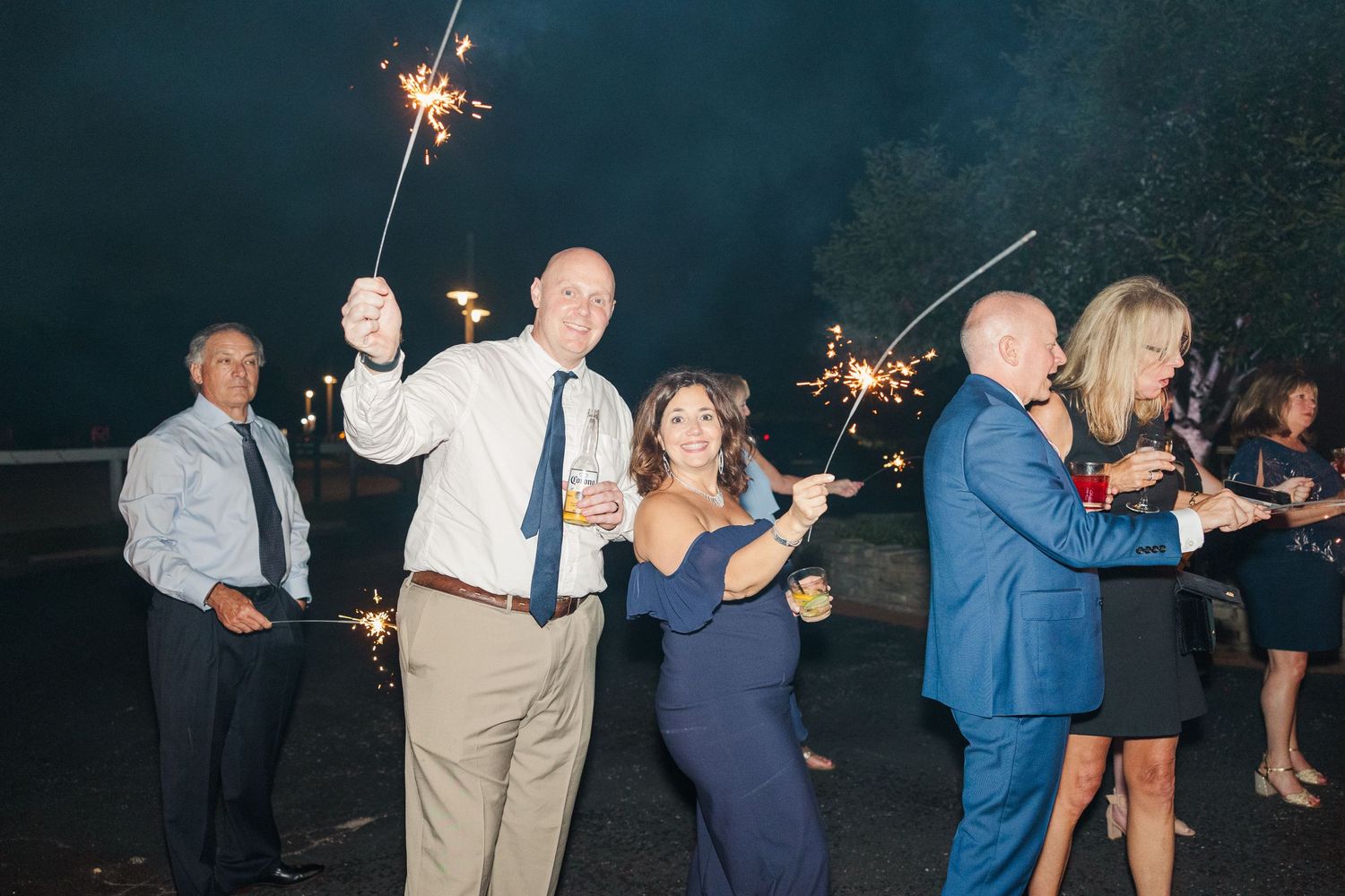 Wedding guests dance and wave sparklers during an outdoor nighttime celebration.
