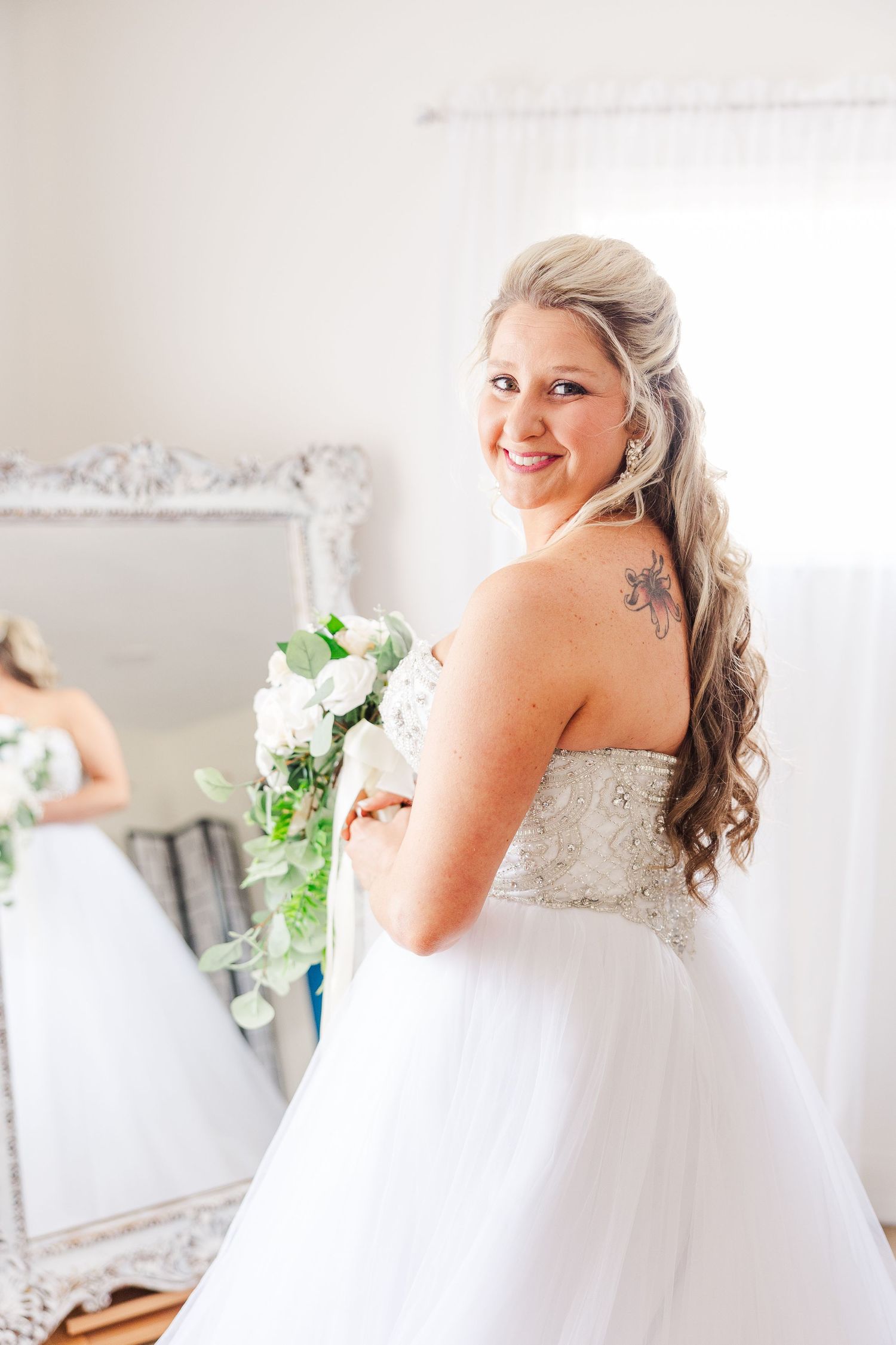Bride in a strapless beaded wedding gown smiles while holding a white bouquet against an ornate mirror background.