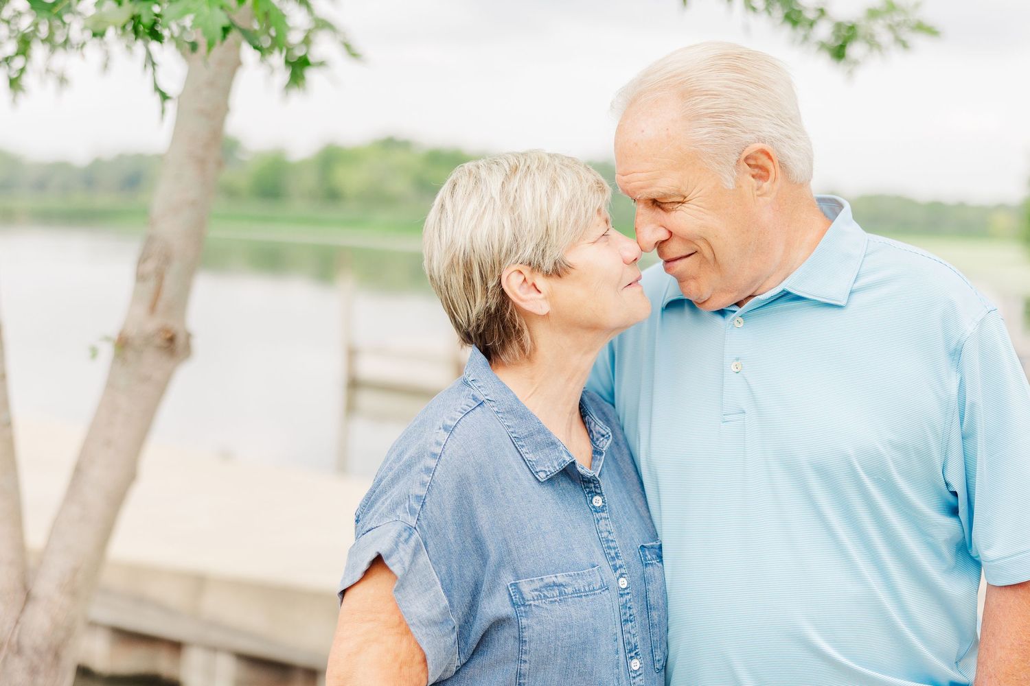 Senior couple sharing an intimate moment together by a lake with trees in the background.