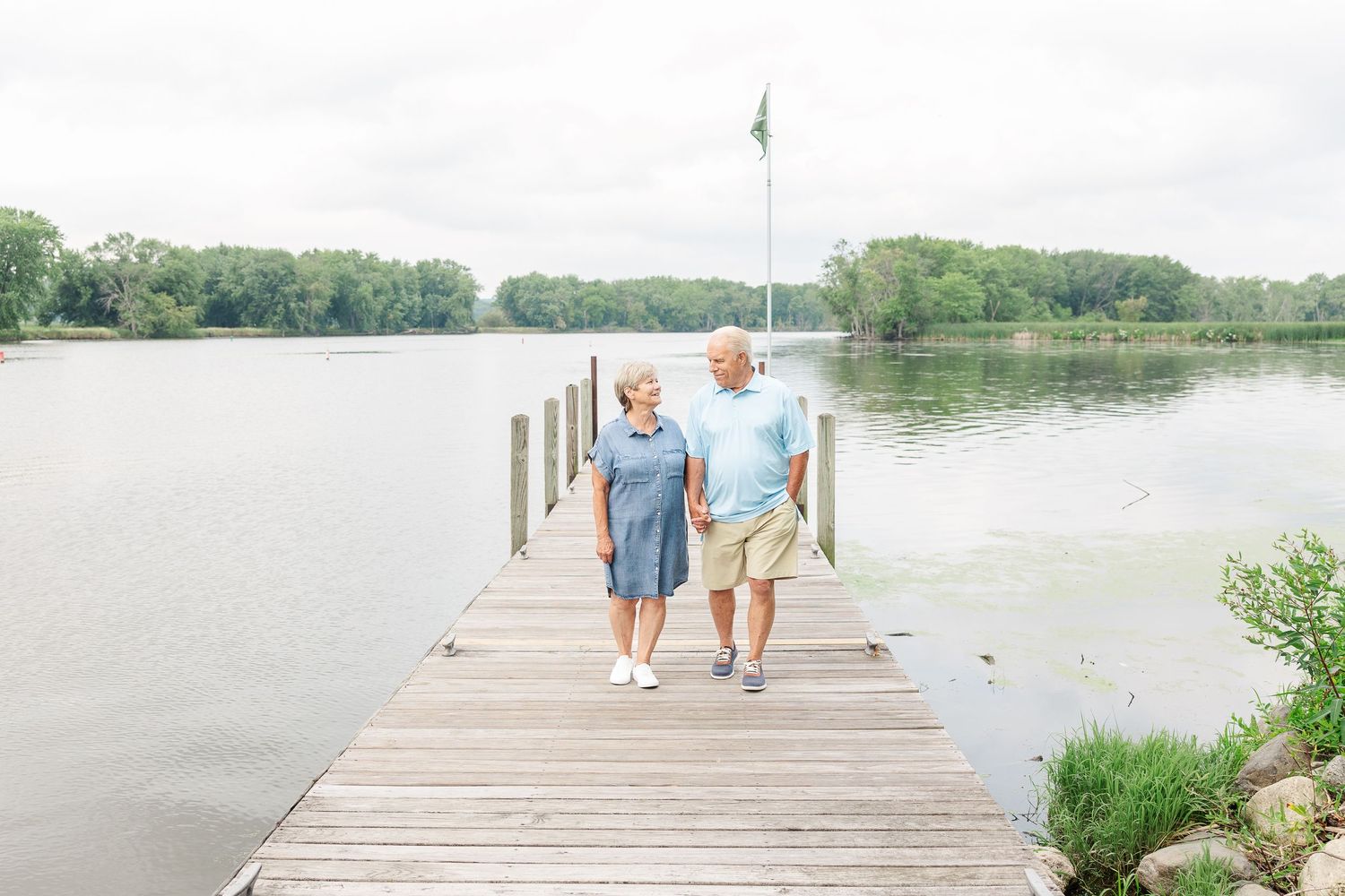 A couple walks hand in hand along a wooden pier overlooking a peaceful lake.