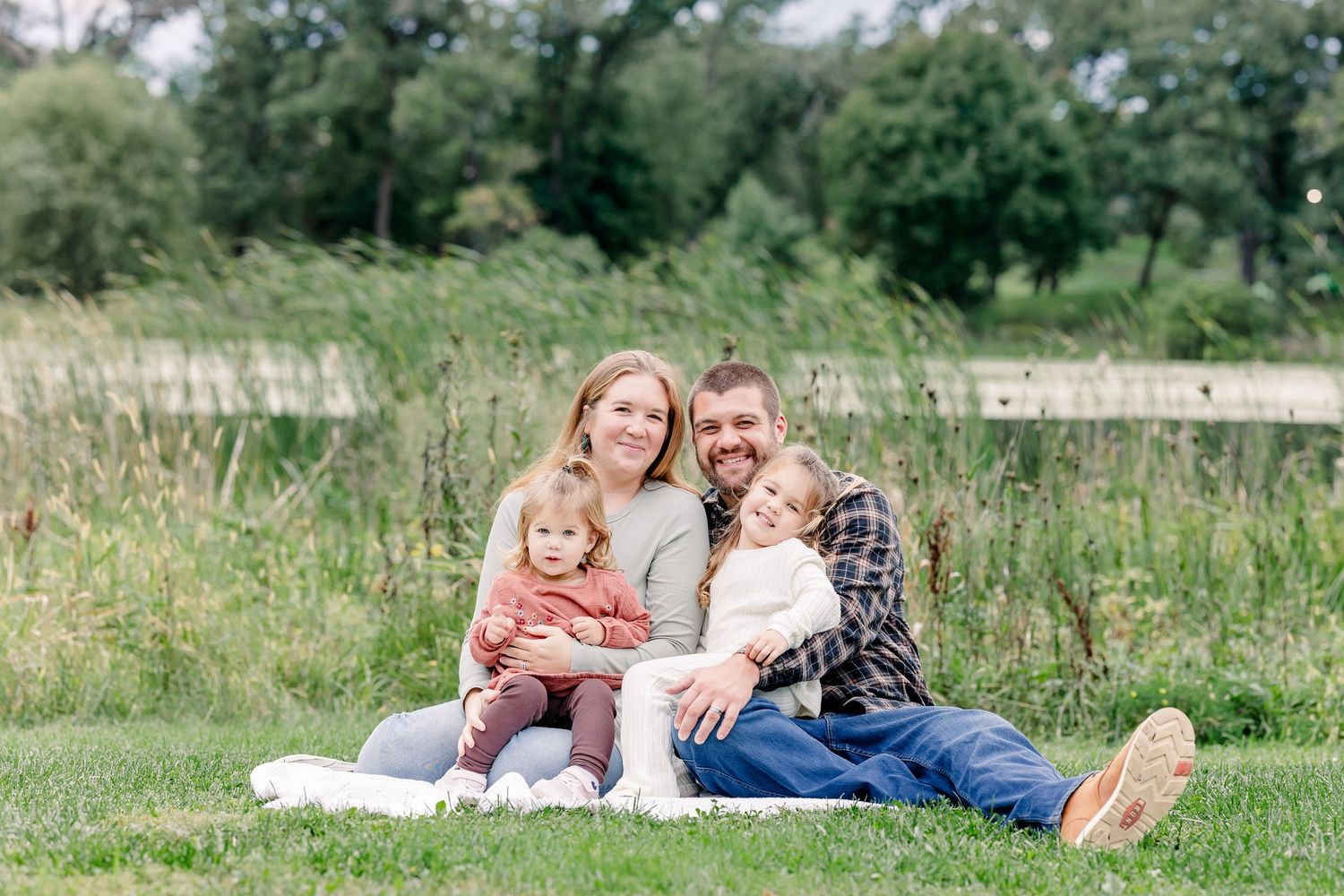 Happy family of four sitting together on green grass near a pond on a sunny day.