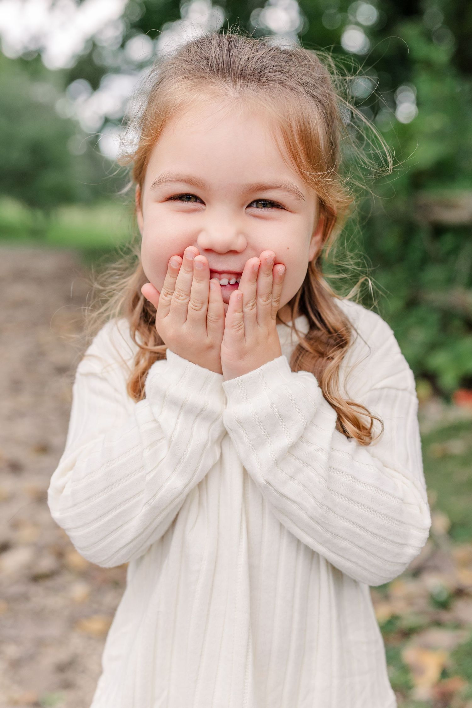 A joyful young girl in a white dress covers her mouth with both hands, smiling outdoors.