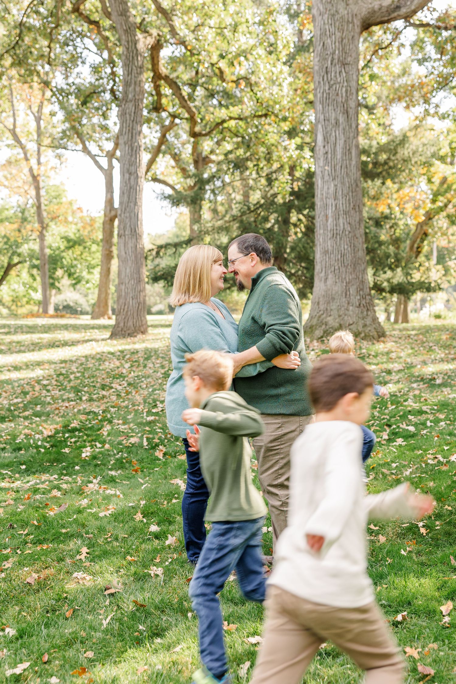 Couple sharing a tender moment nose-to-nose while children run around them in an autumn park.