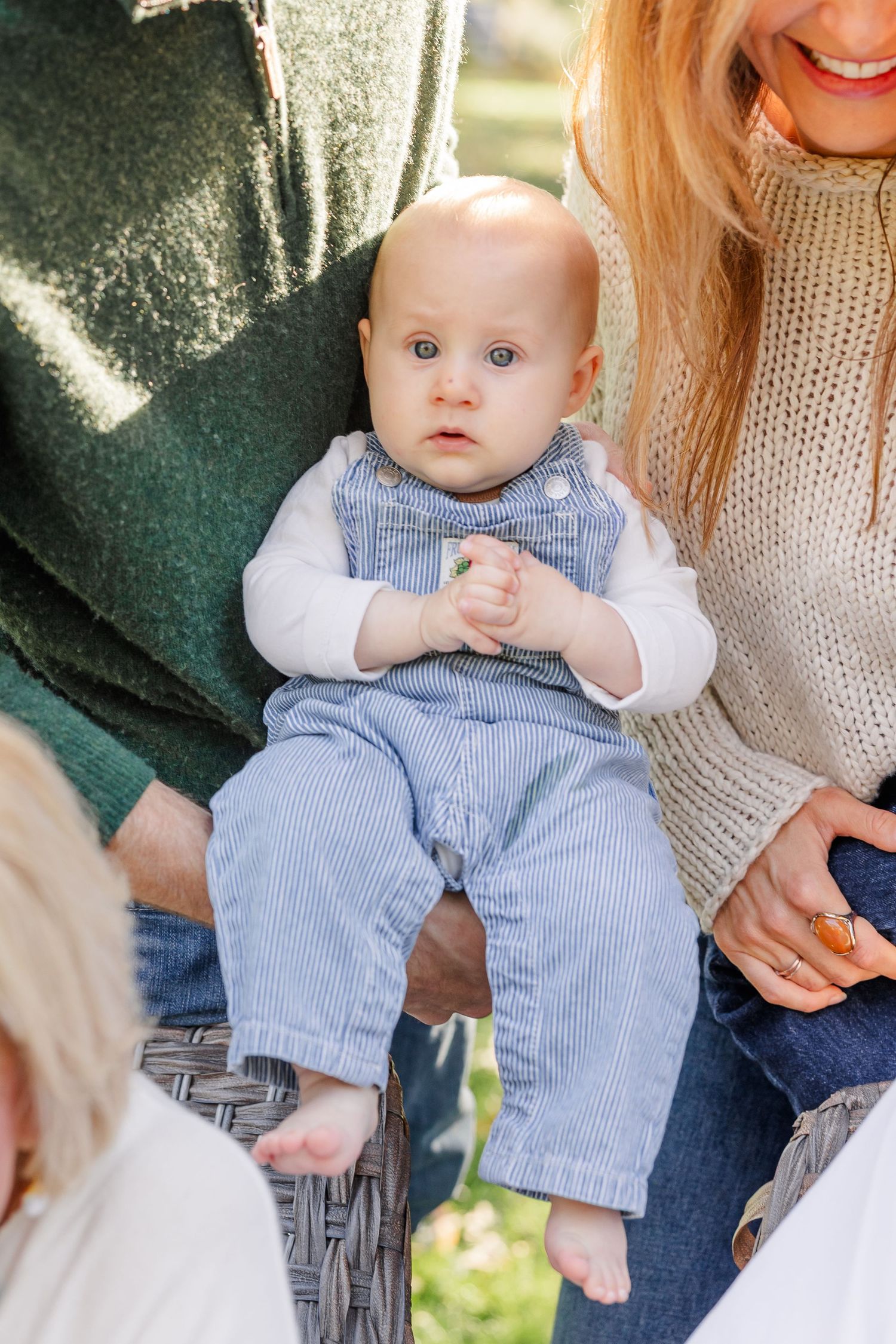Adorable blue-eyed baby in striped overalls sitting between parents outdoors during a family photo session.
