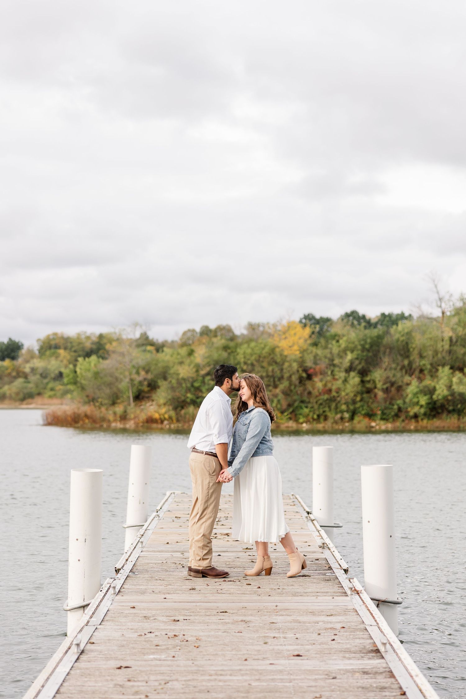 Engaged couple holding hands and kissing at the end of a wooden dock overlooking a calm lake and fall trees under a cloudy sky.