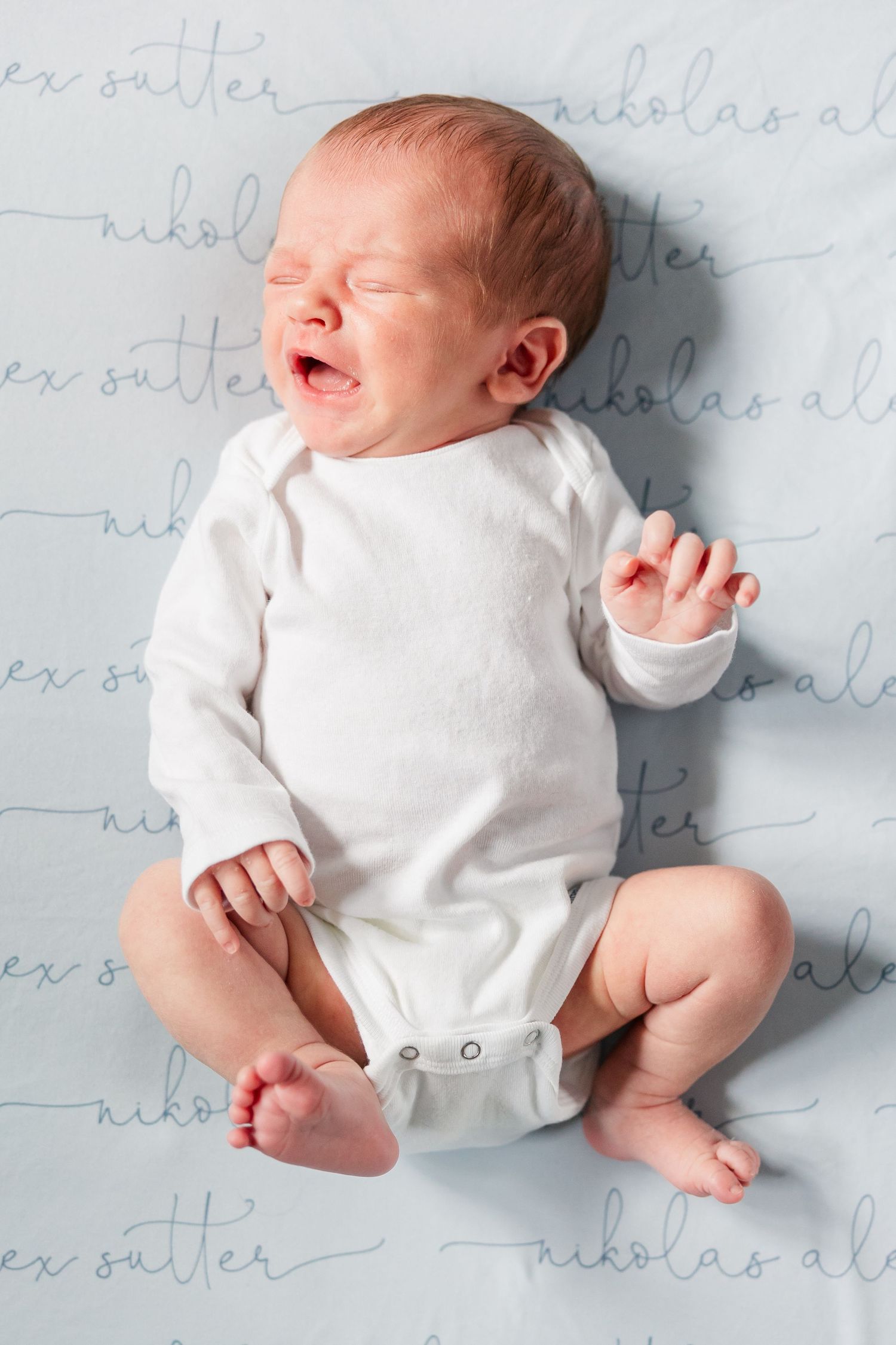 Newborn baby in white onesie crying on personalized blue name blanket reading Nikolas Alex Sutter.