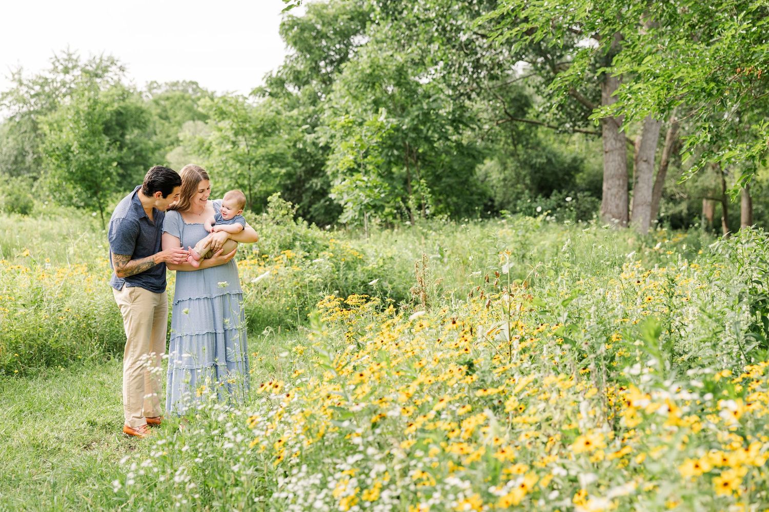 Couple with newborn standing among yellow wildflowers in a lush green park setting.