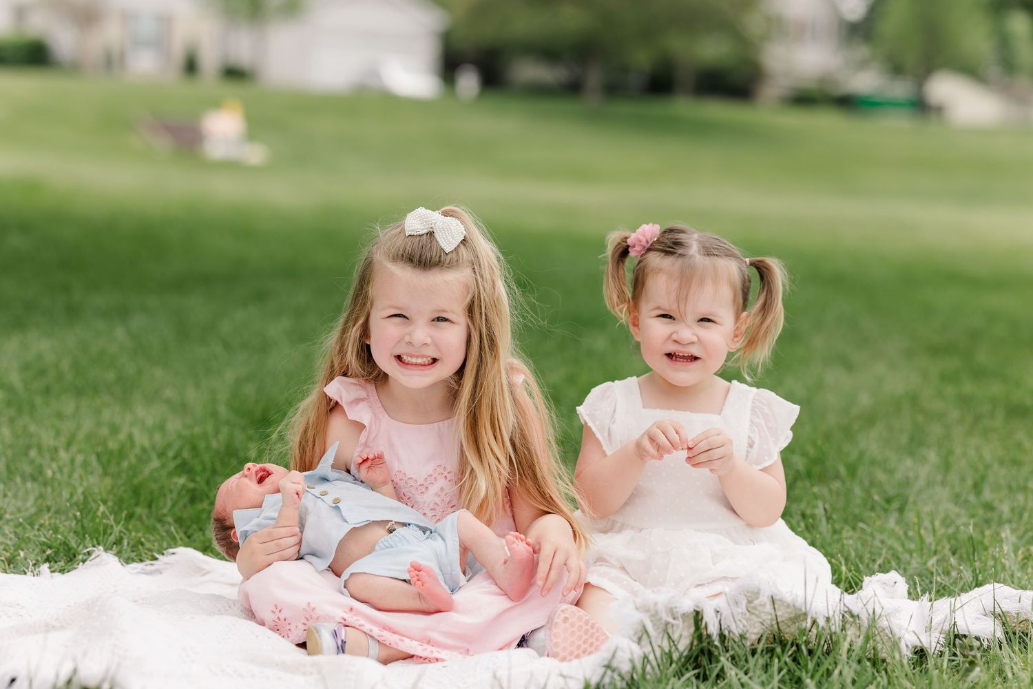 Two smiling young sisters sitting on white blanket in grass holding a newborn baby sibling.