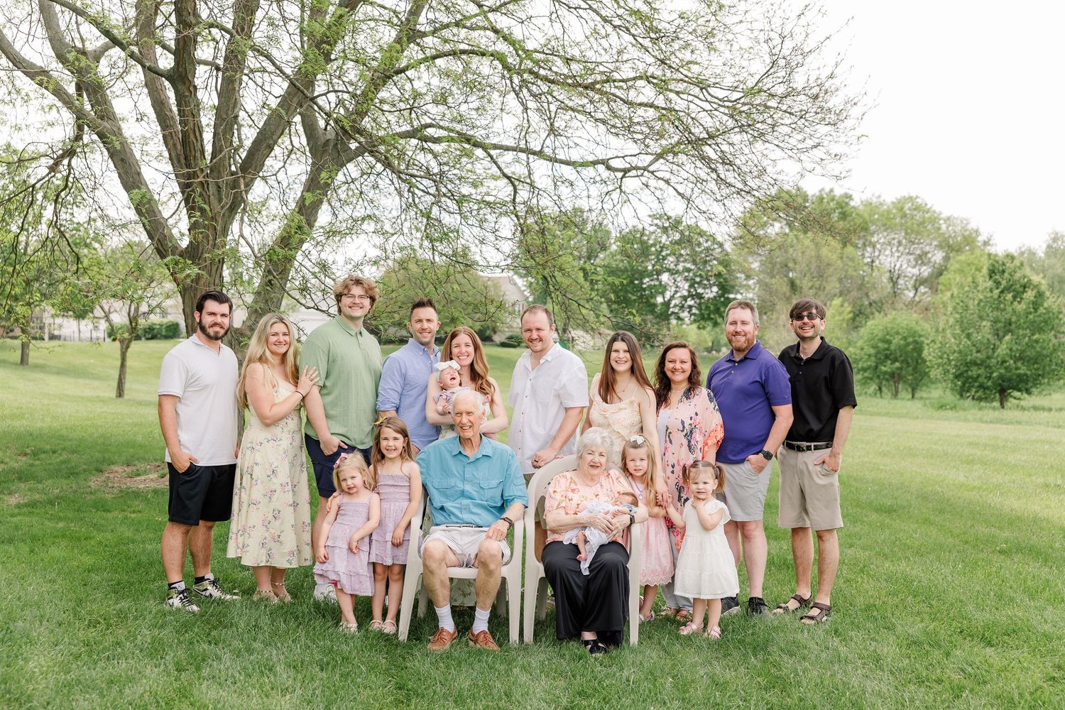 A large multigenerational family poses together outdoors on a green lawn surrounded by spring trees.