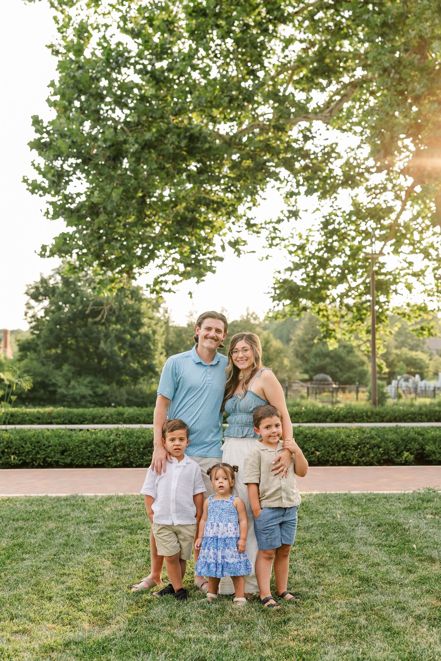 A happy family of five poses on green grass with large trees in a sunny park background.