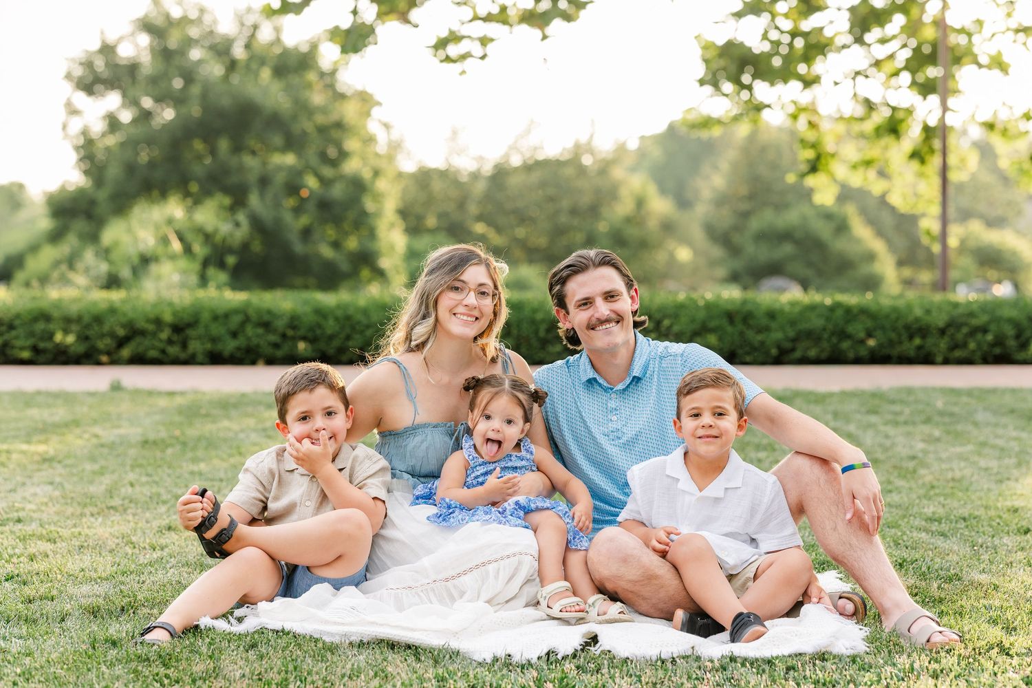 Happy family of five sitting on blanket in sunny park, parents with three young children smiling.