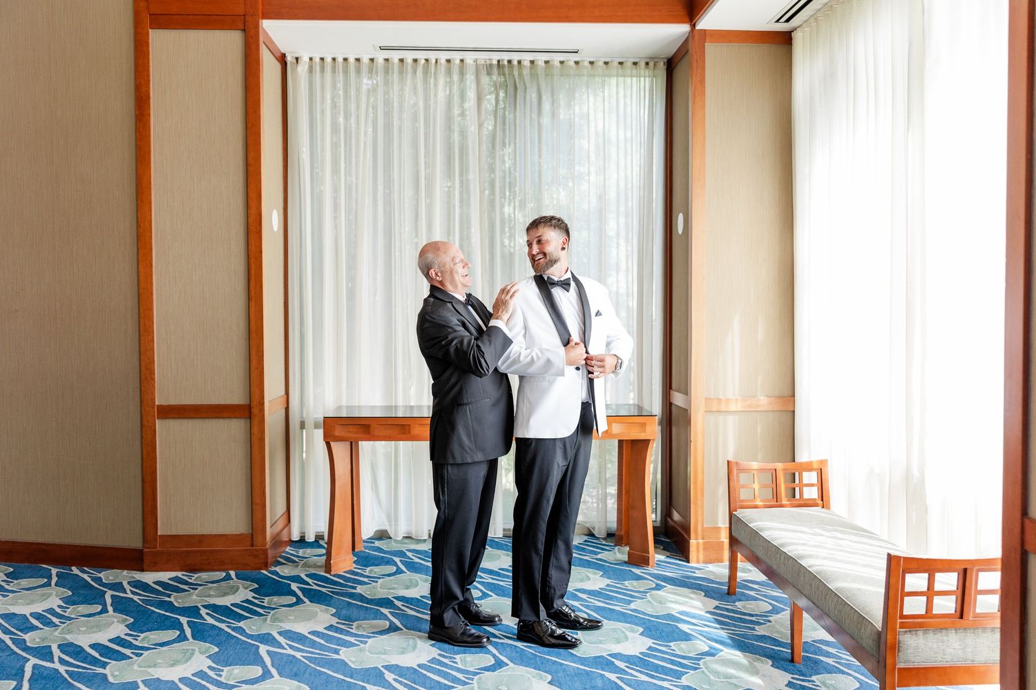 Groom in white tuxedo jacket sharing a moment with older gentleman in black suit indoors.