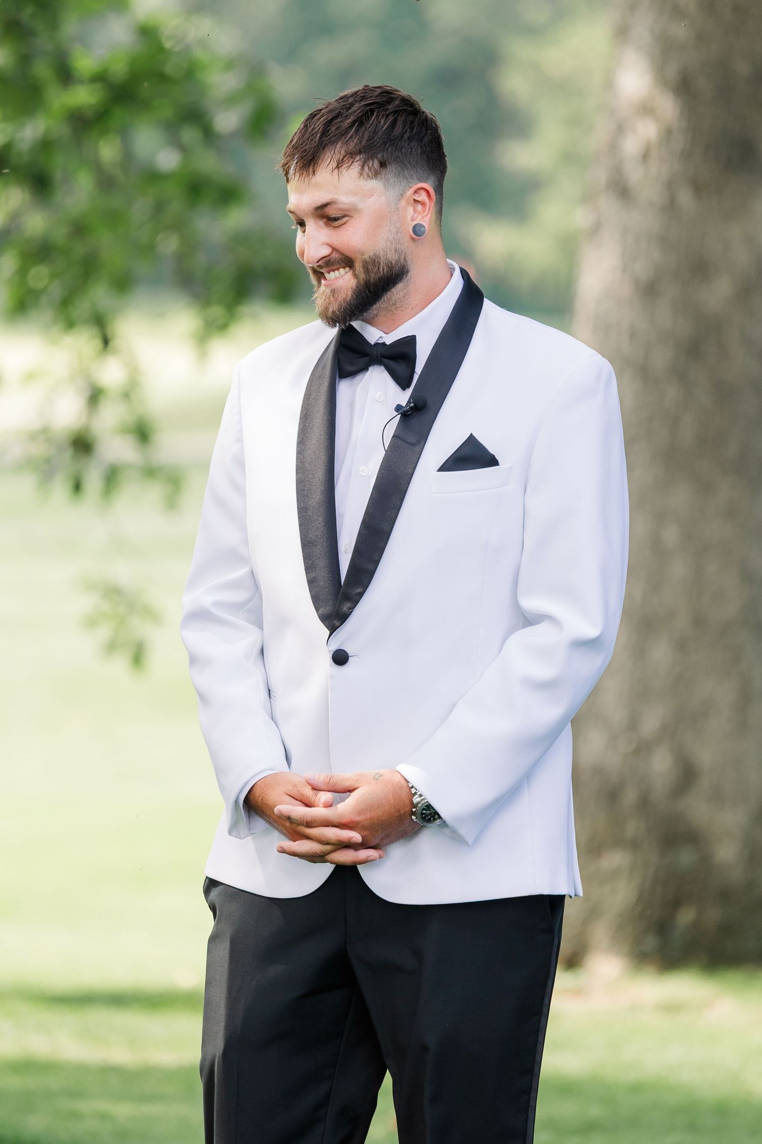 Smiling groom in white tuxedo jacket with black lapels stands outdoors near trees.