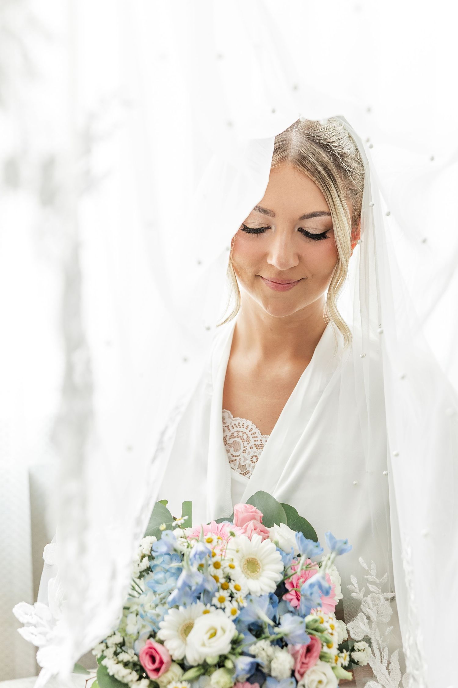 Smiling bride in white veil holds colorful bouquet of pink, blue, and white flowers on wedding day.