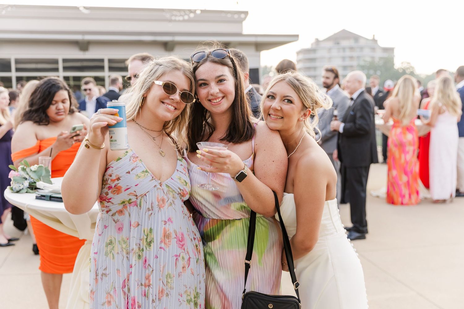 Three smiling women in floral dresses holding drinks at outdoor wedding cocktail reception.