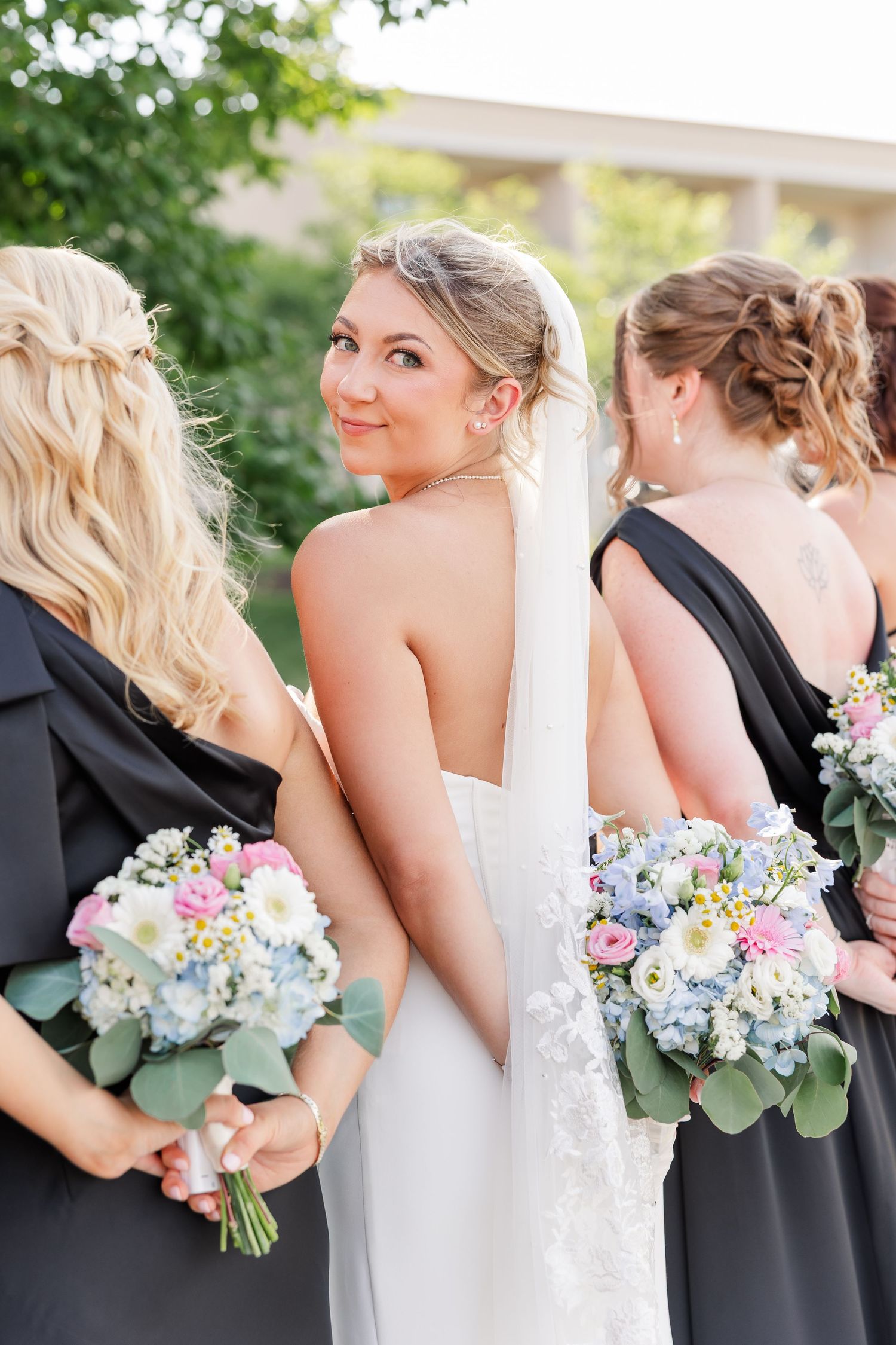 Bride in backless white gown turns to smile while bridesmaids in black dresses hold pastel floral bouquets.