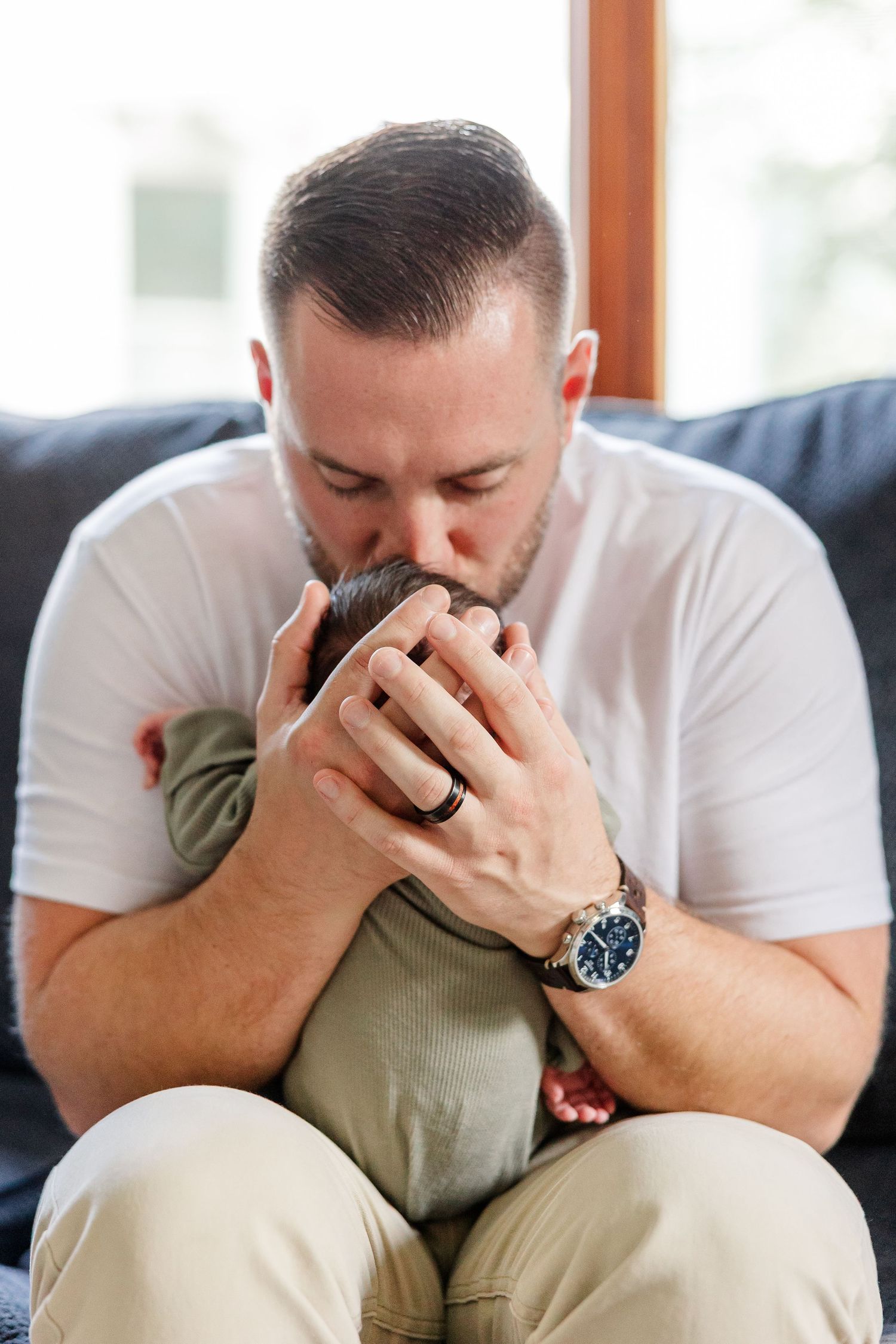 A father tenderly kisses his newborn baby while seated on a dark sofa, wearing a white t-shirt and watch.