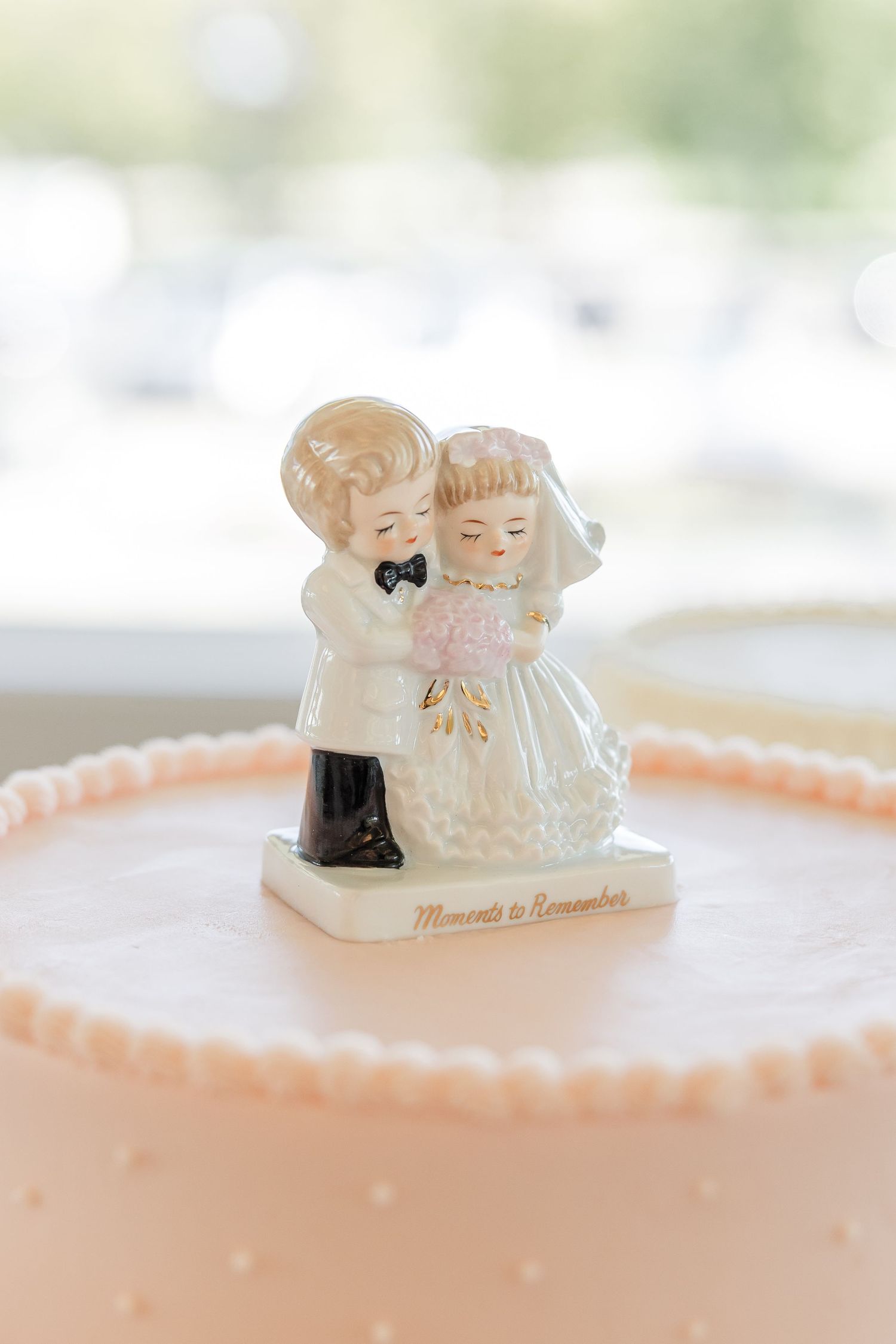 Vintage-style porcelain bride and groom cake topper sits atop a pale pink wedding cake with a pearl-bead border.