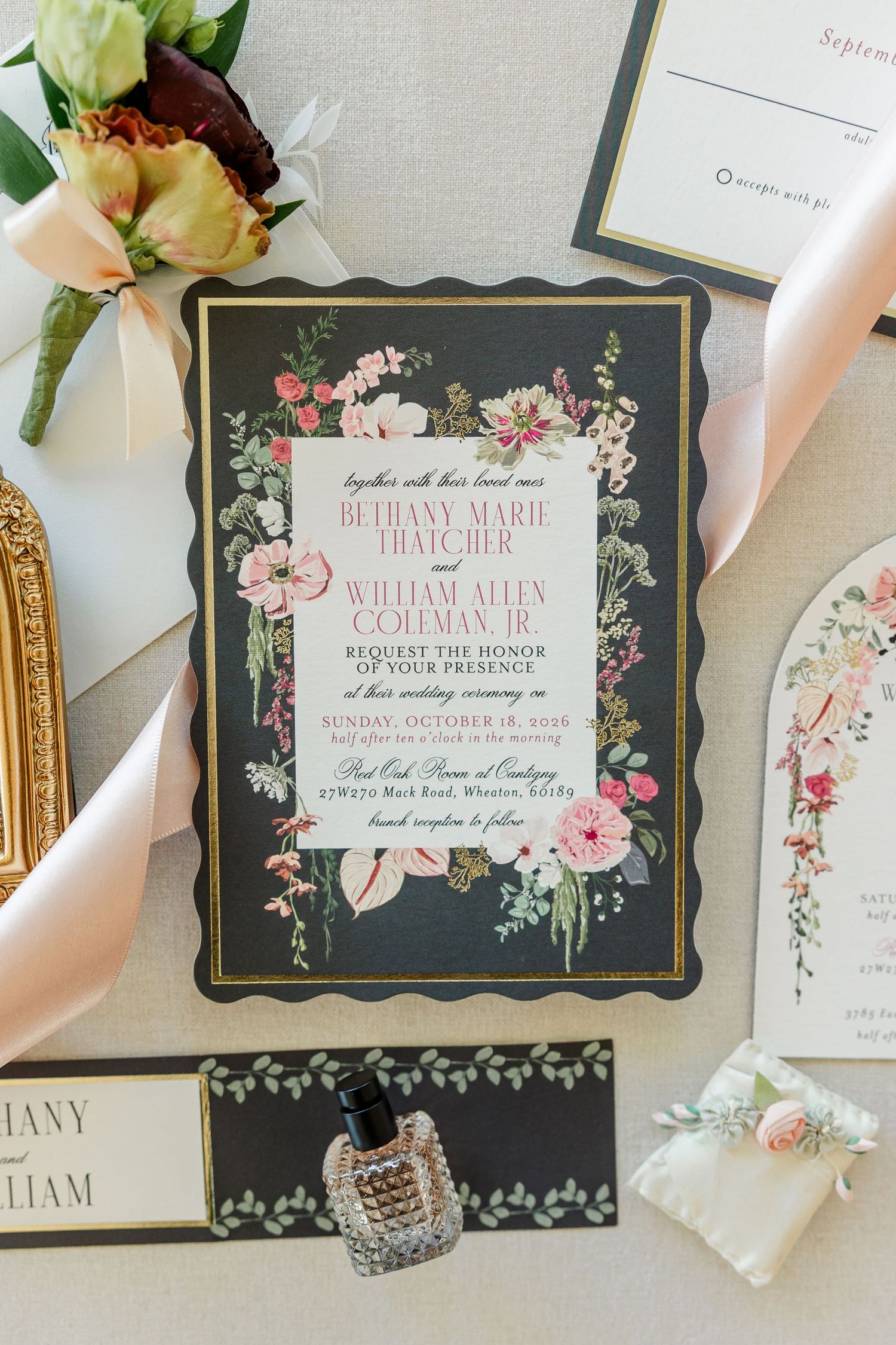 Elegant black and gold wedding invitation with pink floral border, styled with blush ribbon, gold frame, perfume bottle, and a boutonniere.
