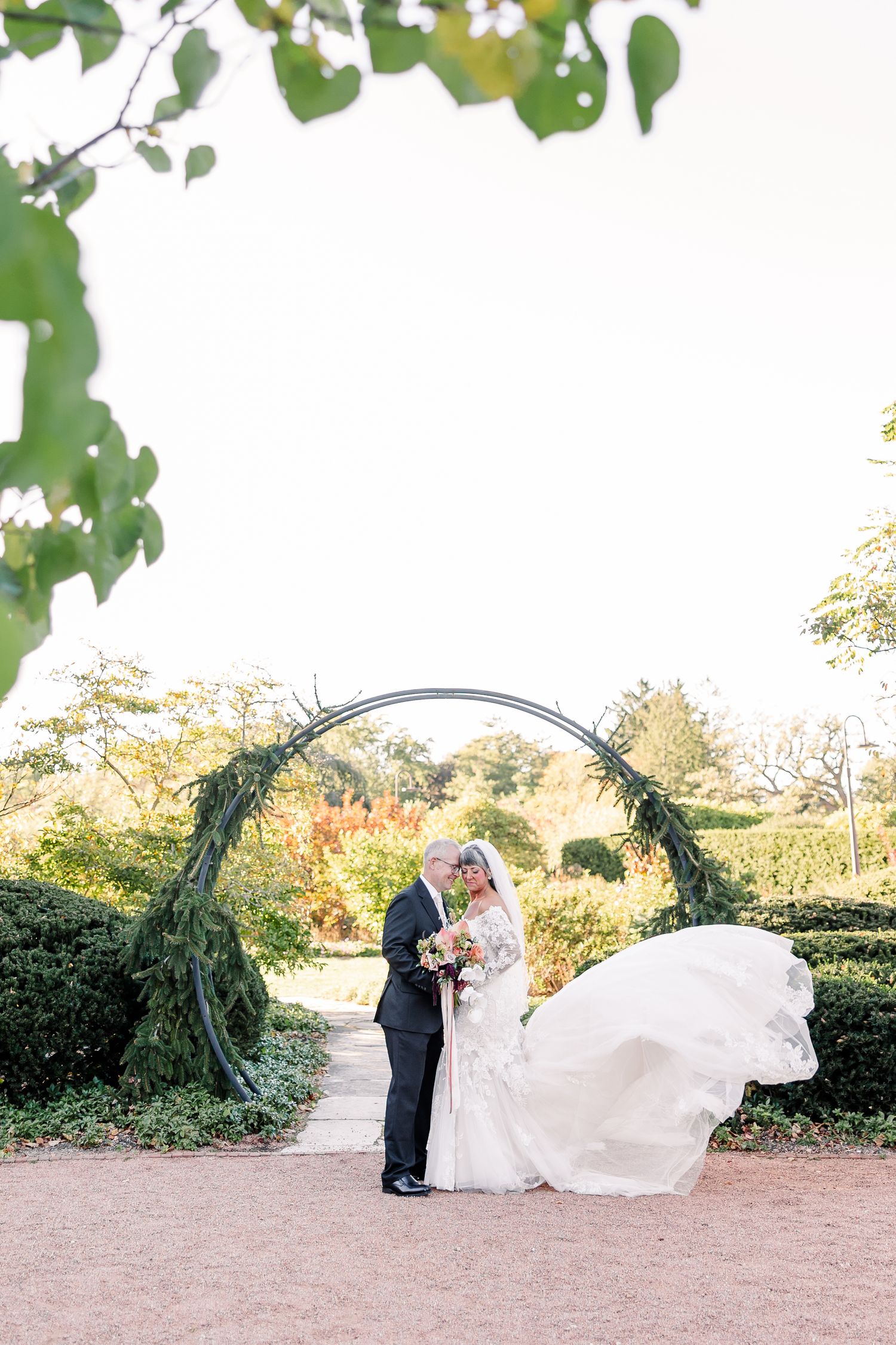 Bride and groom share an intimate moment before a greenery-covered garden arch, her long veil and gown flowing in the breeze.