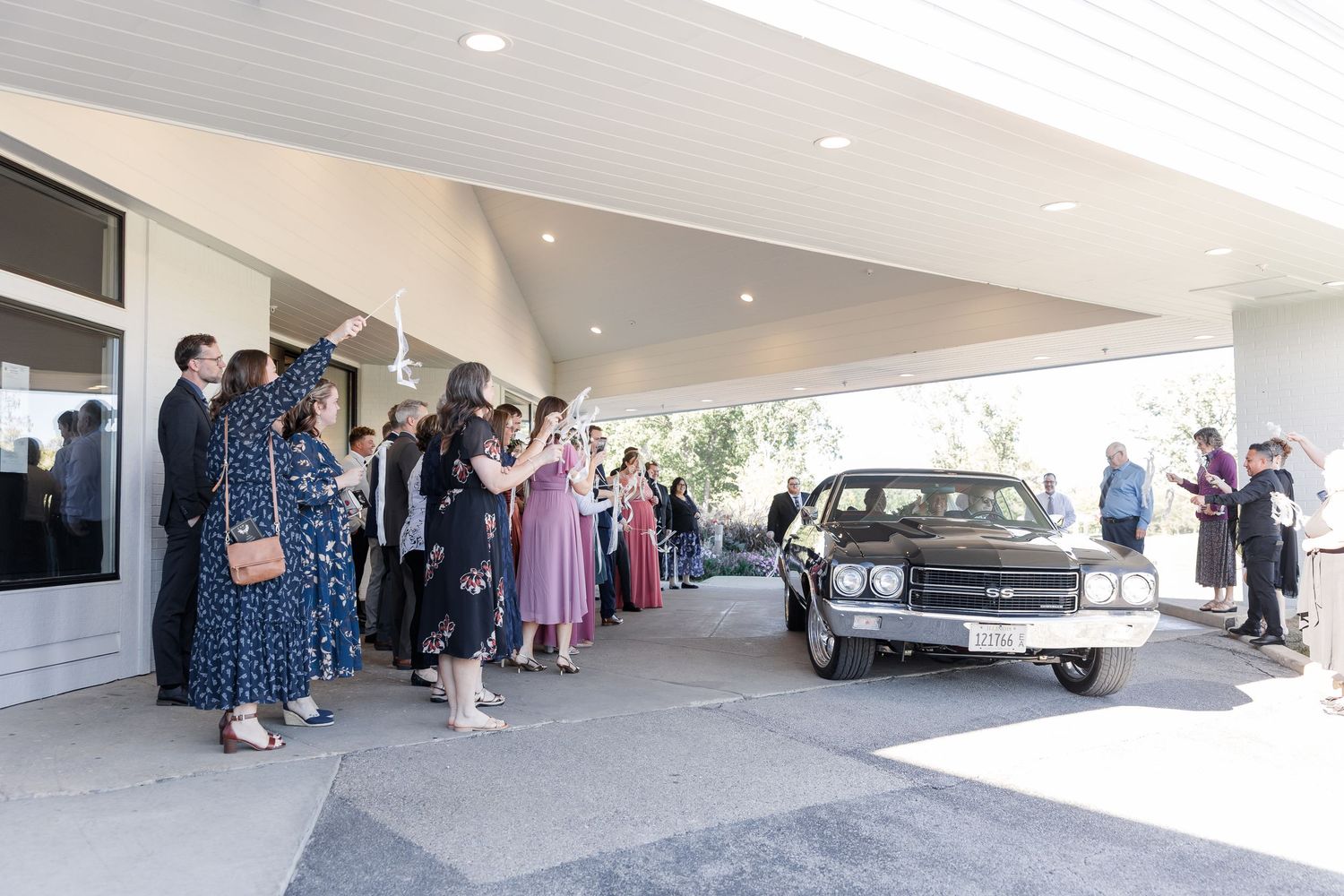 Wedding guests in formal attire cheer and wave as a vintage black car arrives under a covered entrance at the venue.