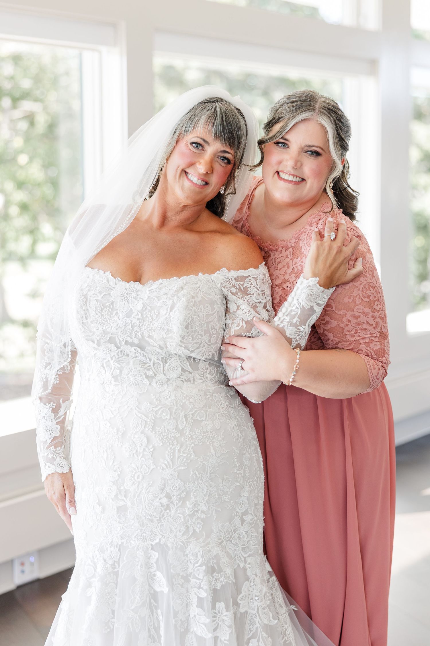 Smiling bride in a lace off-the-shoulder wedding gown and veil embraces a woman in a dusty rose dress beside a bright window.