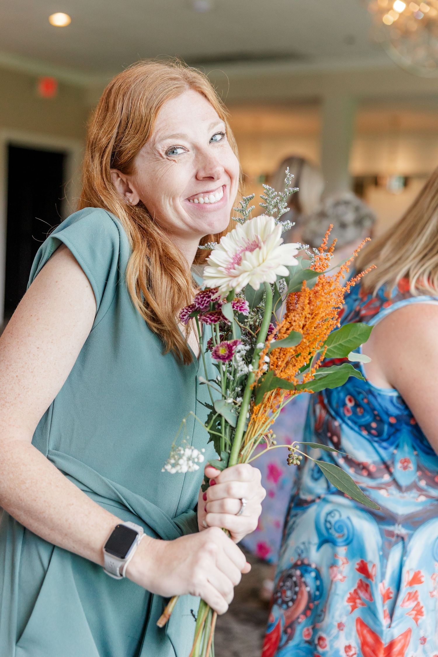 Smiling redhead woman in teal dress holding a wildflower bouquet at an indoor bridal shower event.