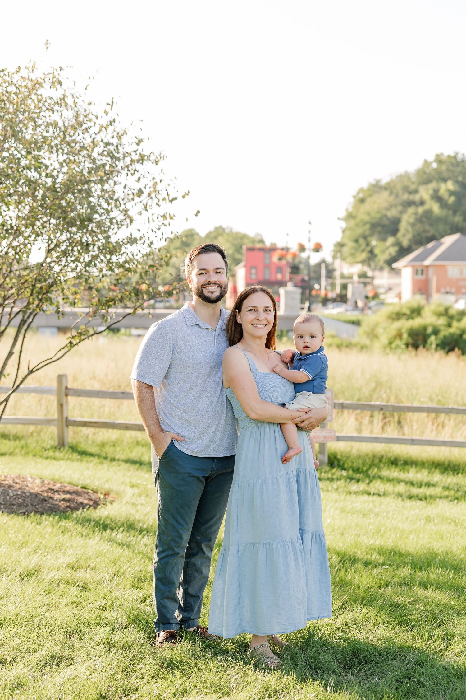 Smiling family of three with dad, mom in blue dress holding baby boy posing in sunny outdoor park.