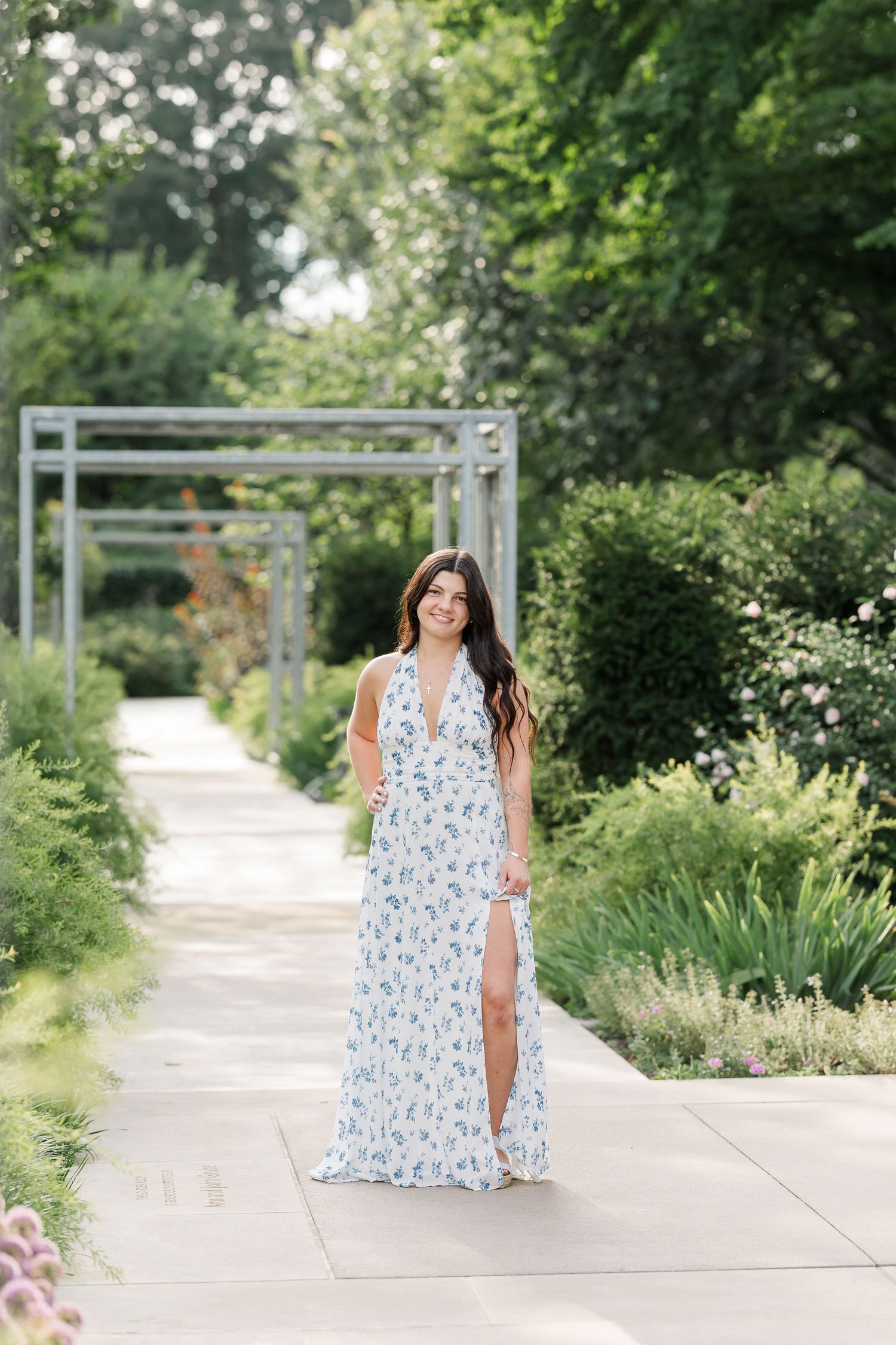 Brunette woman in blue floral maxi dress walking along sunny garden pathway with pergola.