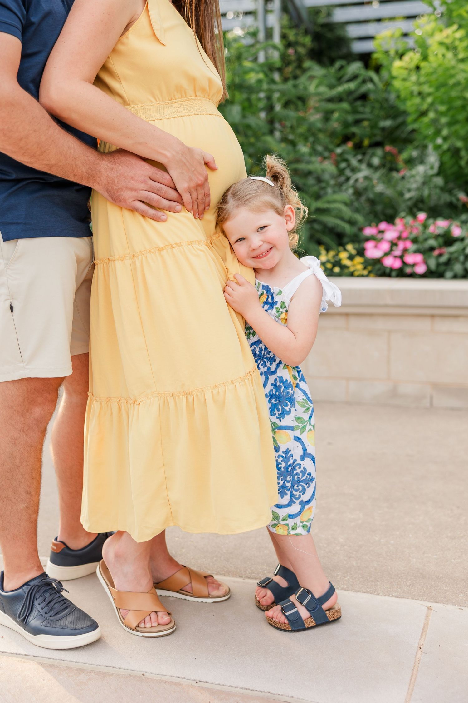 Toddler girl in blue floral dress hugs pregnant mothers yellow dress while father stands beside them outdoors.