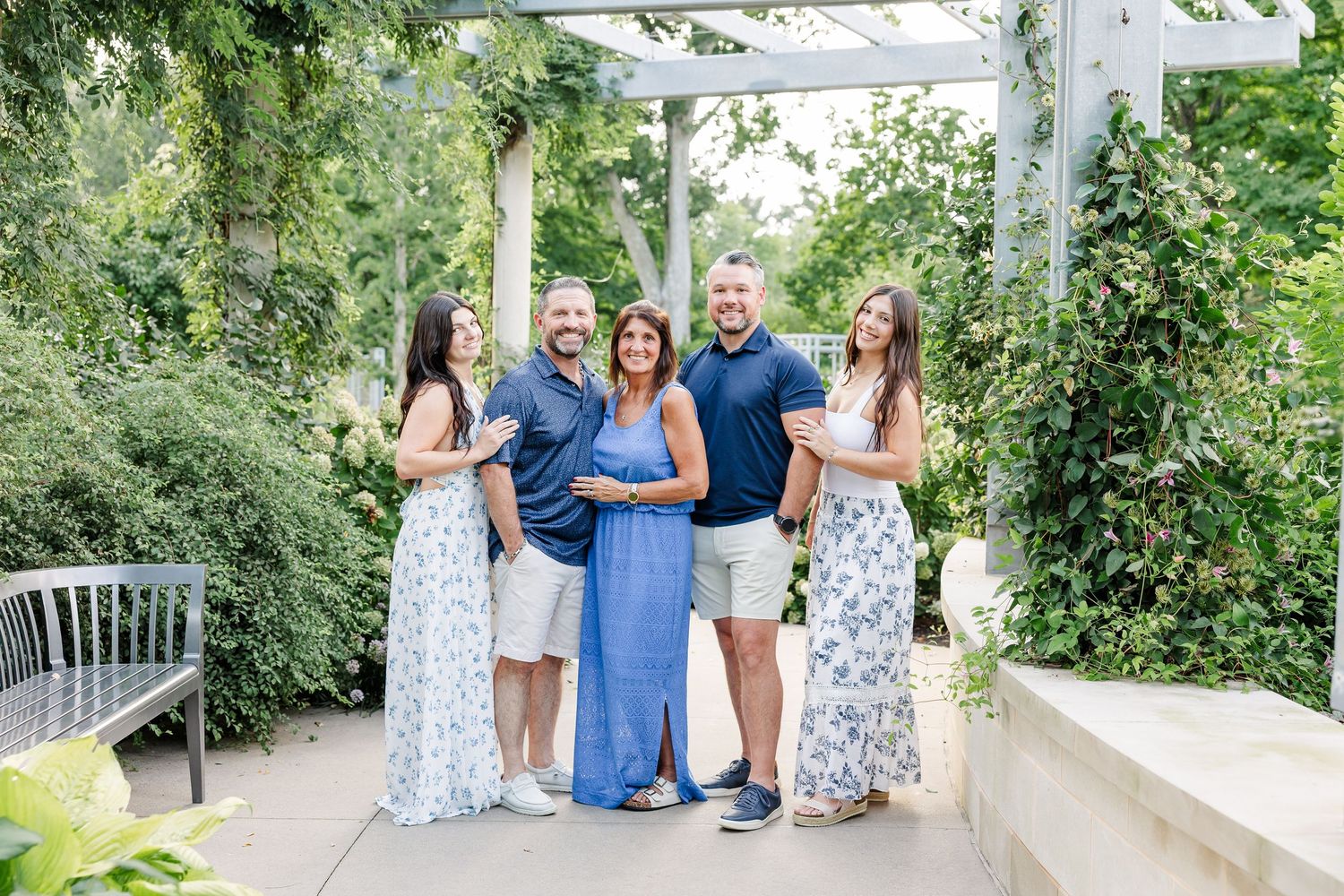 Family of five smiling together under a vine-covered pergola in a beautiful garden setting.
