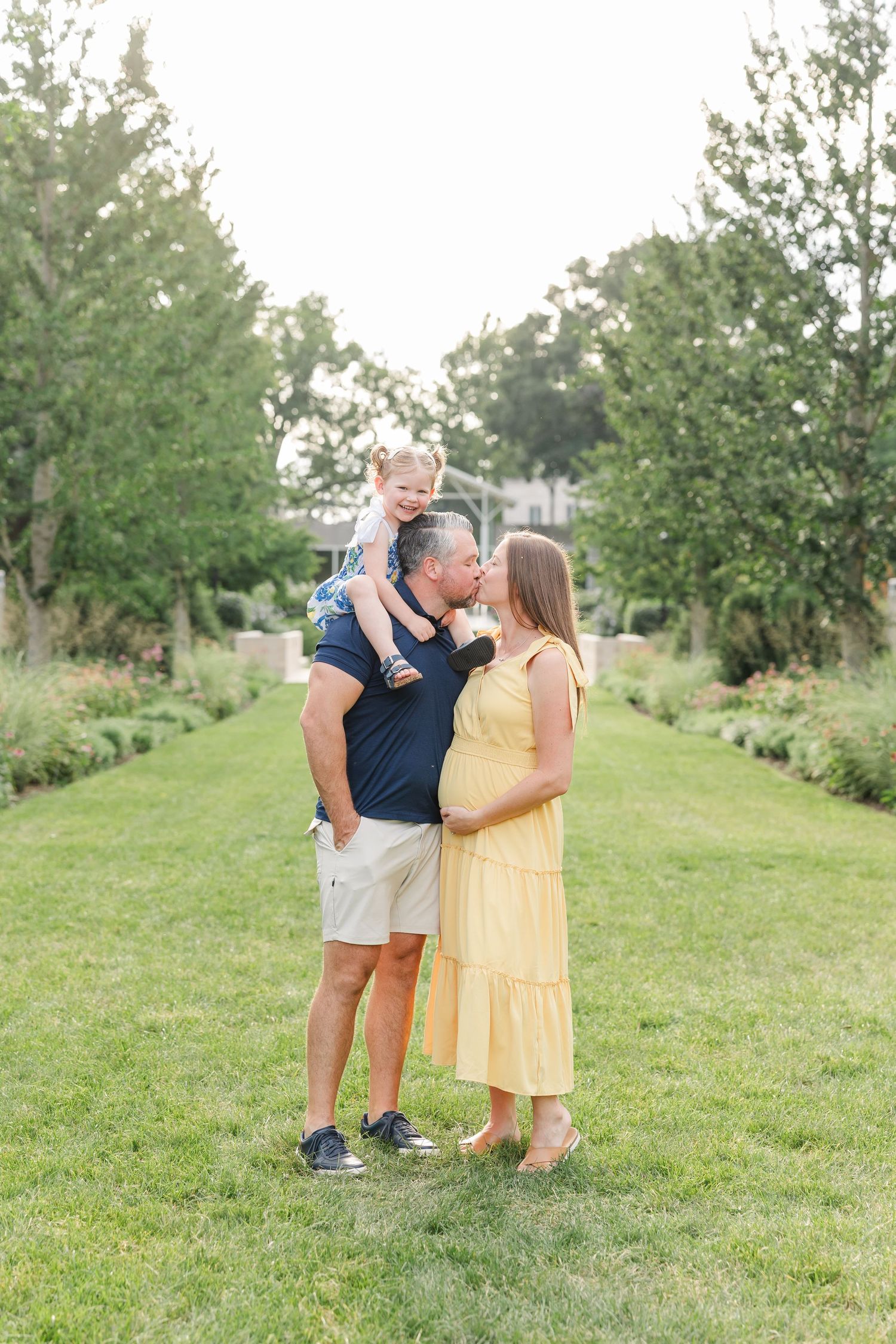 A pregnant woman in a yellow dress and a man in navy holding a toddler share a kiss outdoors in a lush garden.