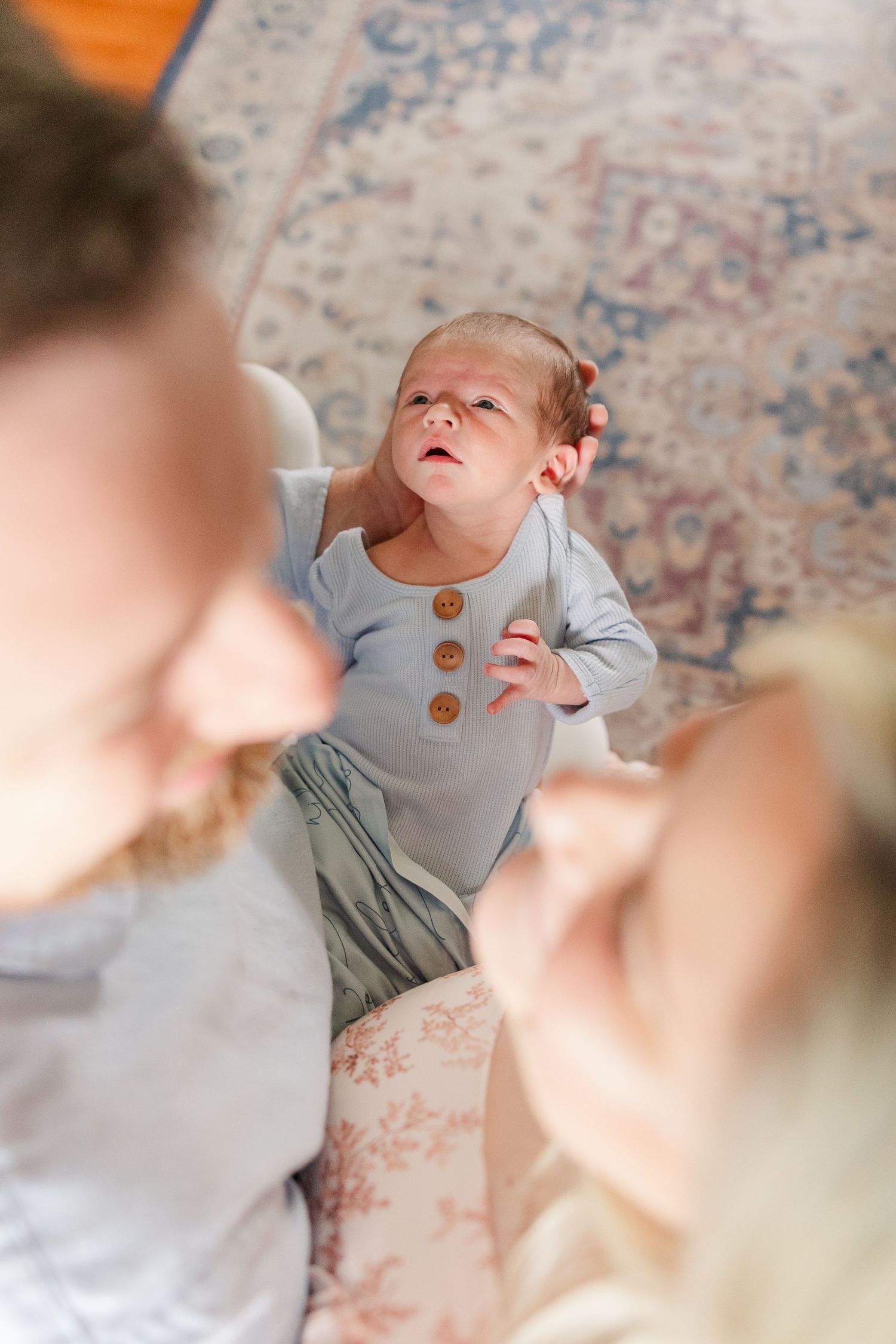 Newborn baby in blue onesie looking up at parents from above in a cozy home setting.