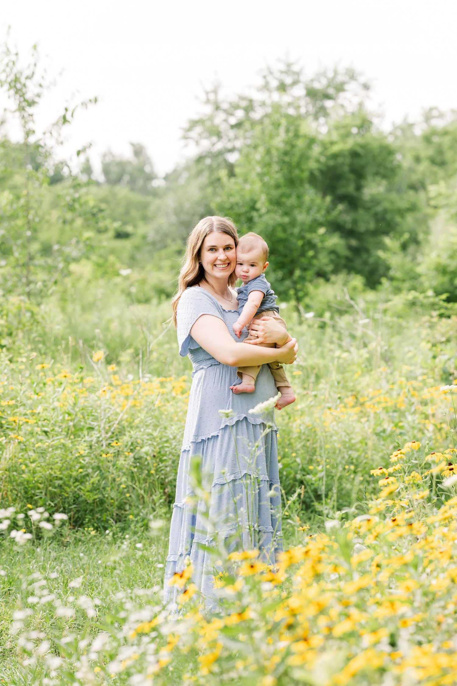 A smiling mother in a blue dress holds her baby in a vibrant yellow wildflower field outdoors.