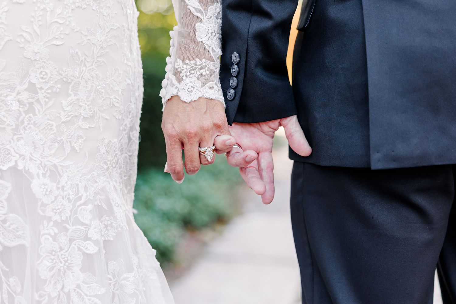 Bride in a white lace wedding dress holding hands with groom in a dark suit, wedding rings visible, outdoors near greenery.