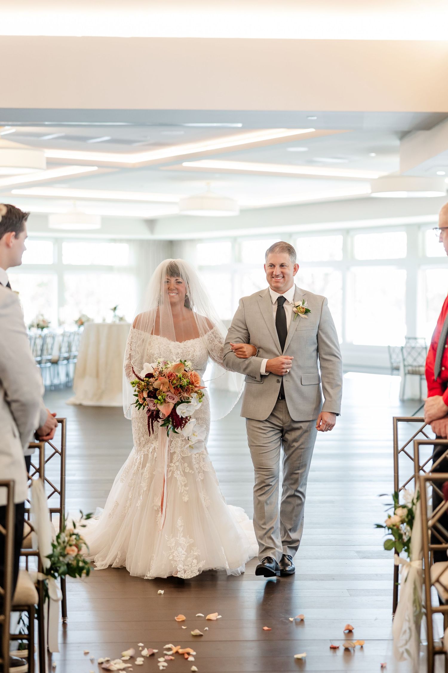 Smiling bride holding a pink and peach bouquet walks arm in arm with a man in a gray suit down a petal-strewn aisle.