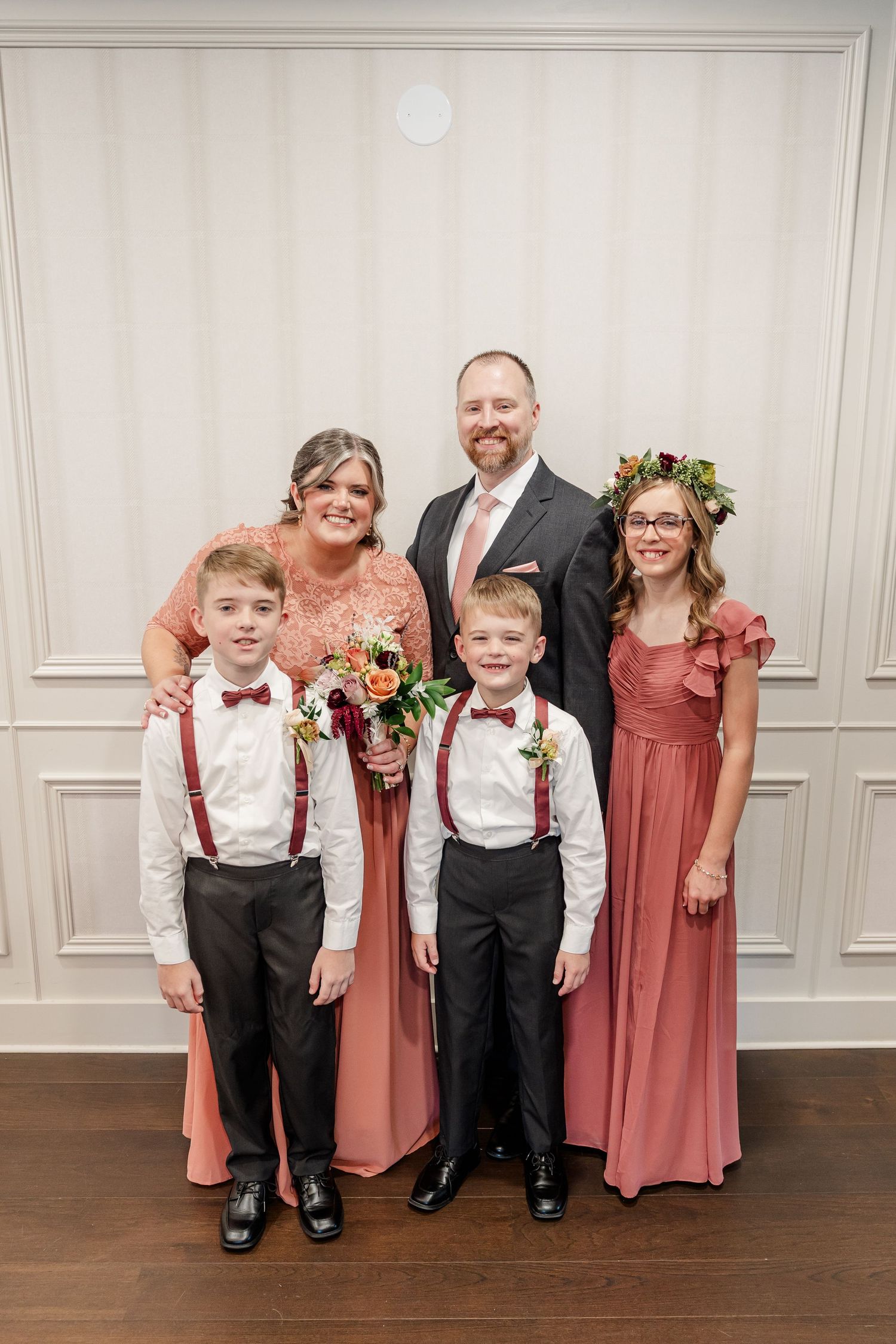 Family of five in coral and blush wedding attire poses together, with two boys in suspenders and bow ties and a girl in a flower crown.
