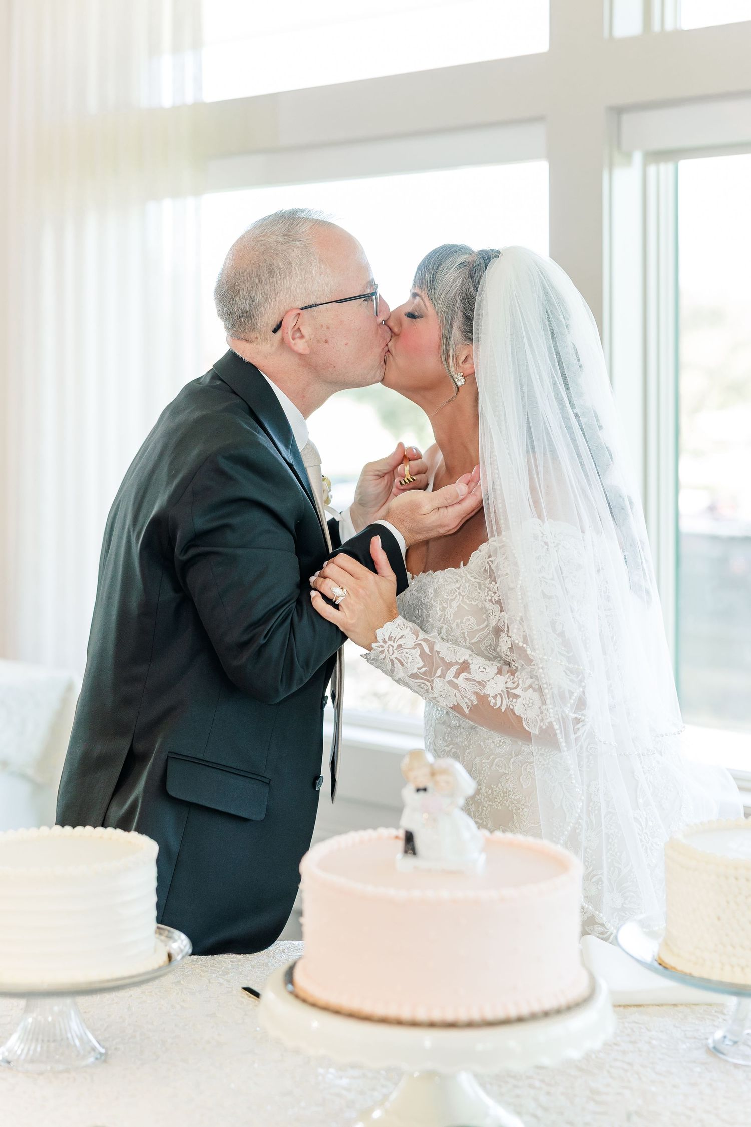 Bride in a veil and lace gown shares a kiss with her groom beside a pink wedding cake topped with a couple figurine.